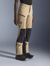 Vrouw draagt Alpinestars Stella Flex-AST Explorer Pants, zandkleurige en zwarte adventure motorbroek met zwarte ripstop kniepanelen, cargozakken met ritssluiting, 'ALL TERRA' logo patch en ritsen aan de onderkant van de pijpen.
