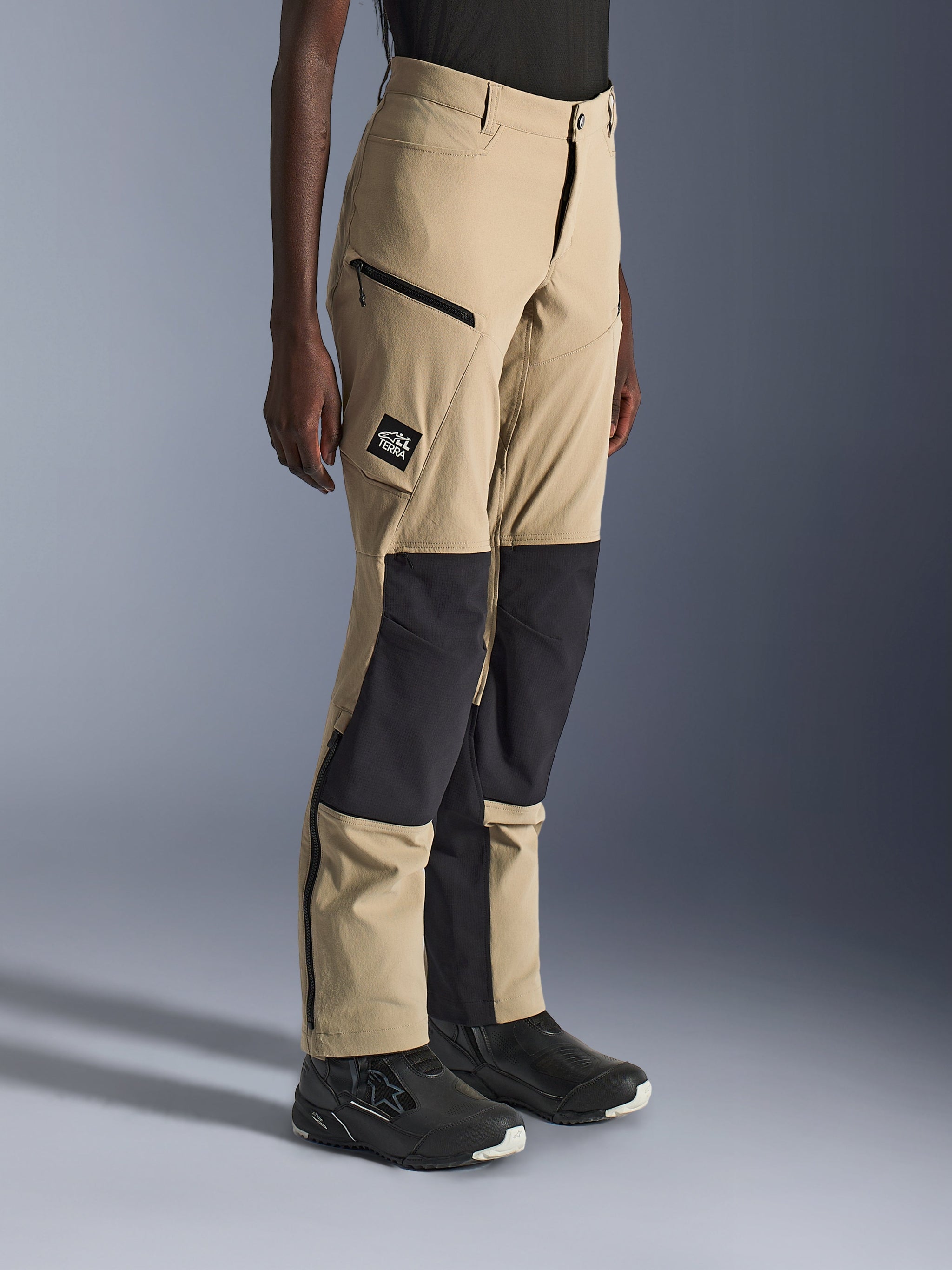Vrouw draagt Alpinestars Stella Flex-AST Explorer Pants, zandkleurige en zwarte adventure motorbroek met zwarte ripstop kniepanelen, cargozakken met ritssluiting, 'ALL TERRA' logo patch en ritsen aan de onderkant van de pijpen.