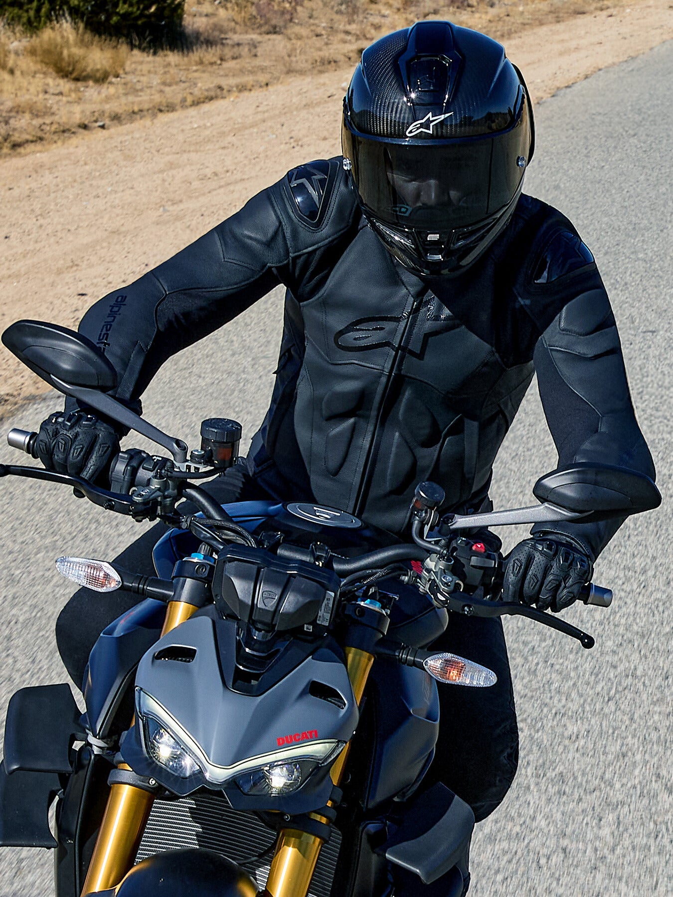 Motorrijder op een grijze Ducati, die de Alpinestars Dusk Leather Jacket in het zwart draagt, voorzien van logo's in reliëf en externe schouderbescherming, gecombineerd met een zwarte integraalhelm en motorhandschoenen voor een premium, op de weg gerichte look.