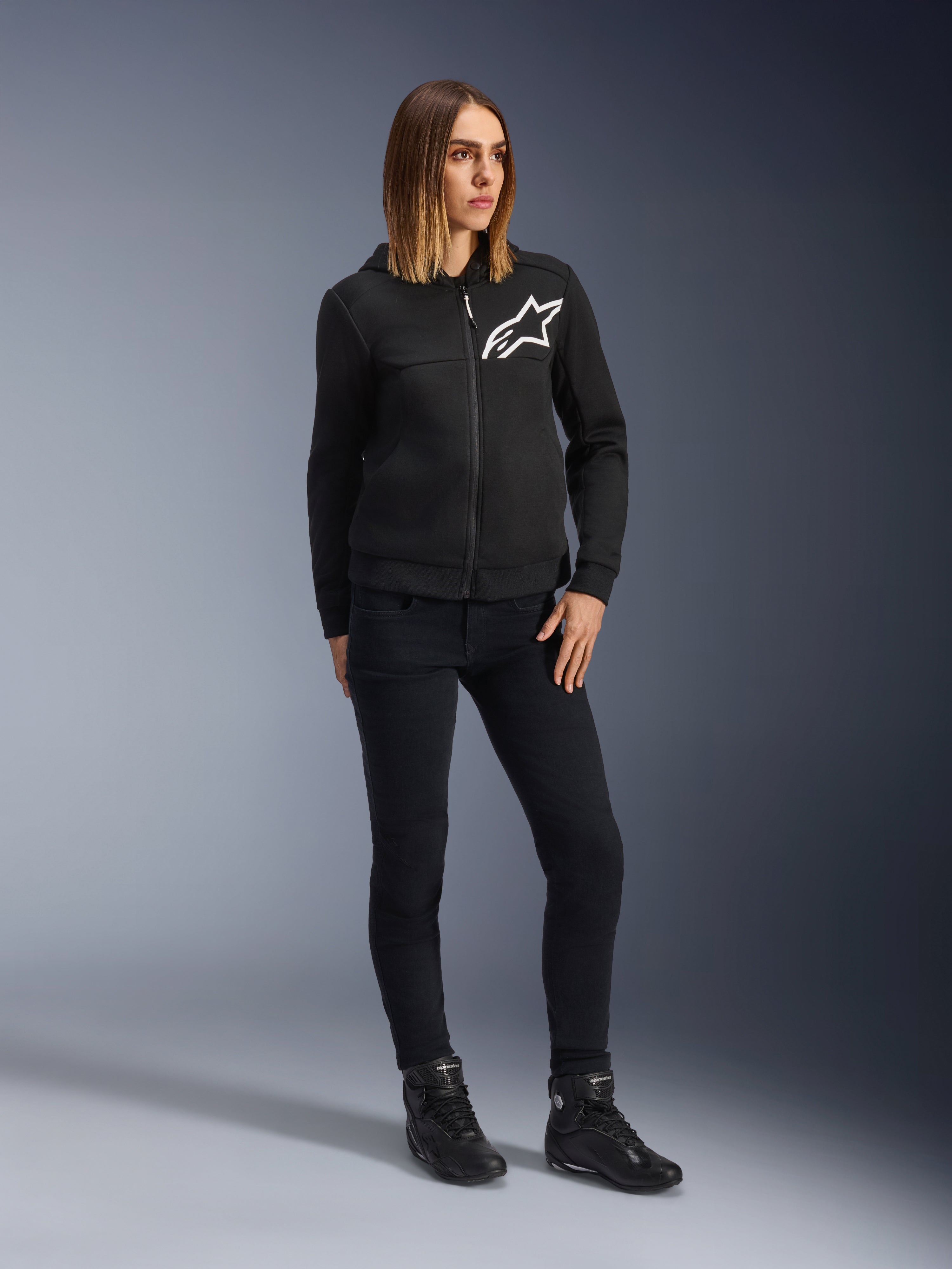 Vrouw draagt Alpinestars Daisy V3 Riding Denim voor dames, slim-fit motorbroek in Rinse Black, gecombineerd met een zwarte hoodie met ritssluiting en motorlaarzen, urban casual beschermende kleding