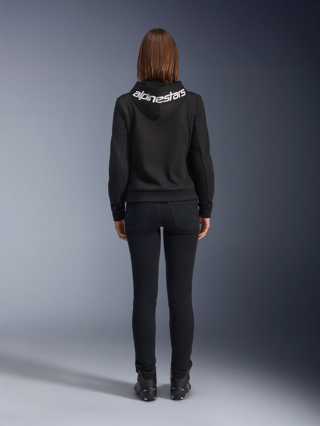 Alpinestars Daisy V3 dames motorjeans, Black Rinse, slim fit motorbroek, achteraanzicht met hoge taille en vrouwelijk silhouet, gecombineerd met een zwarte Alpinestars hoodie en zwarte motorlaarzen, urban casual motorkleding