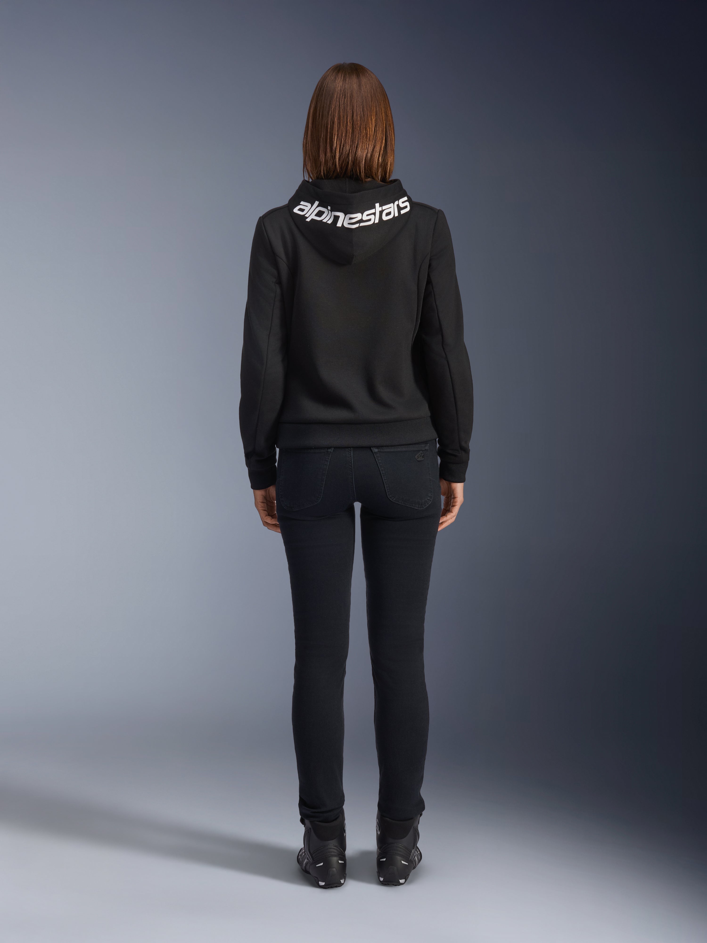 Alpinestars Daisy V3 dames motorjeans, Black Rinse, slim fit motorbroek, achteraanzicht met hoge taille en vrouwelijk silhouet, gecombineerd met een zwarte Alpinestars hoodie en zwarte motorlaarzen, urban casual motorkleding