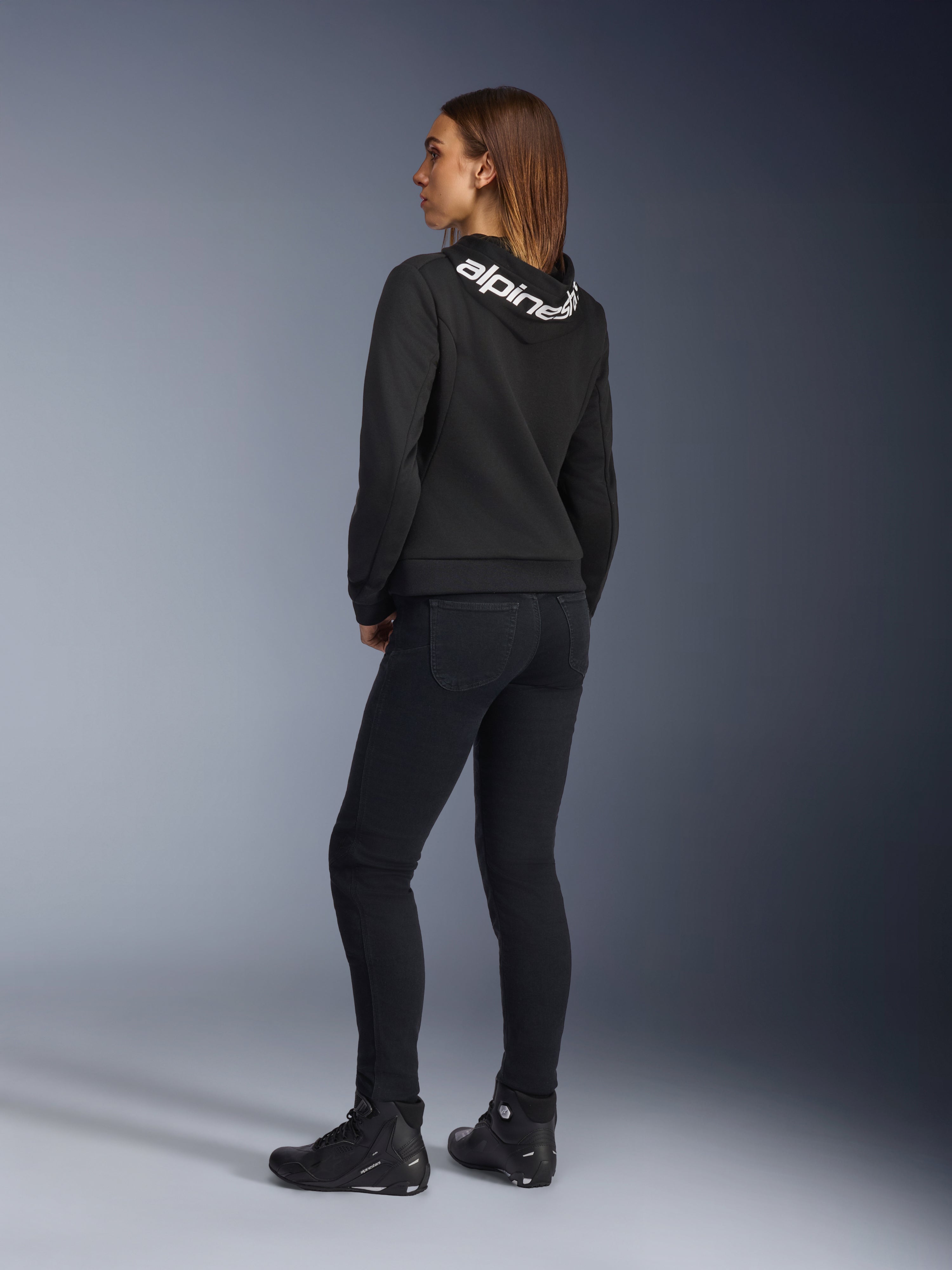 Alpinestars Daisy V3 dames motorjeans, motorbroek voor dames, Rinse Black, achteraanzicht met slanke pasvorm en hoge taille, gecombineerd met een zwarte Alpinestars hoodie en motorschoenen voor een urban motorlook