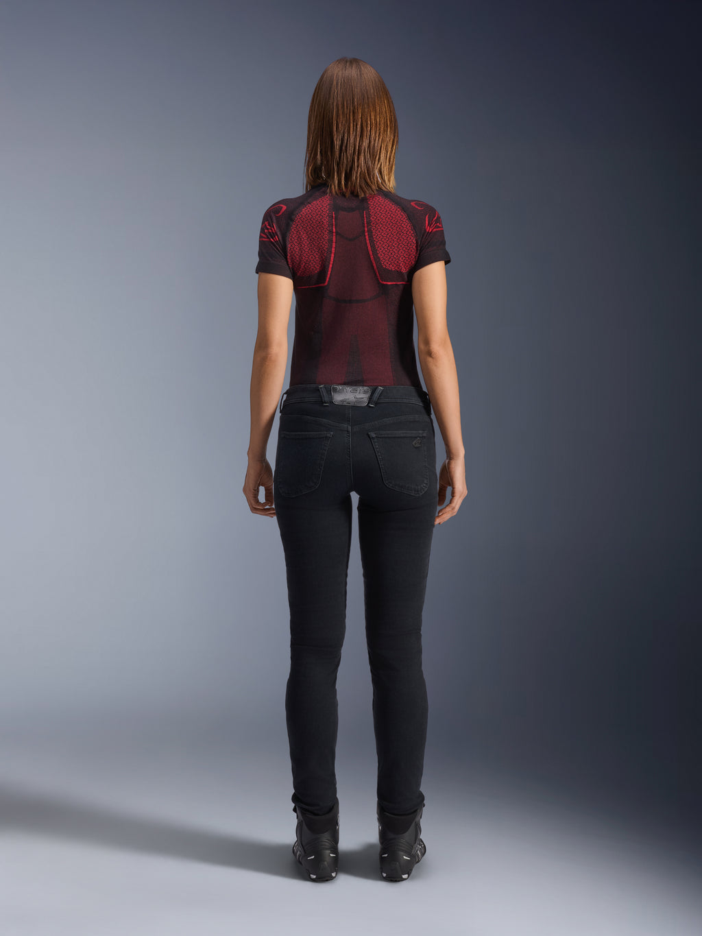 Alpinestars Daisy V3 dames motorjeans, Rinse Black, slim-fit motorbroek van achteren gefotografeerd, met een high-waist ontwerp en achterzakken, gecombineerd met een rood en zwart technisch shirt met patroon en zwarte laarzen, urban motorkleding voor dames