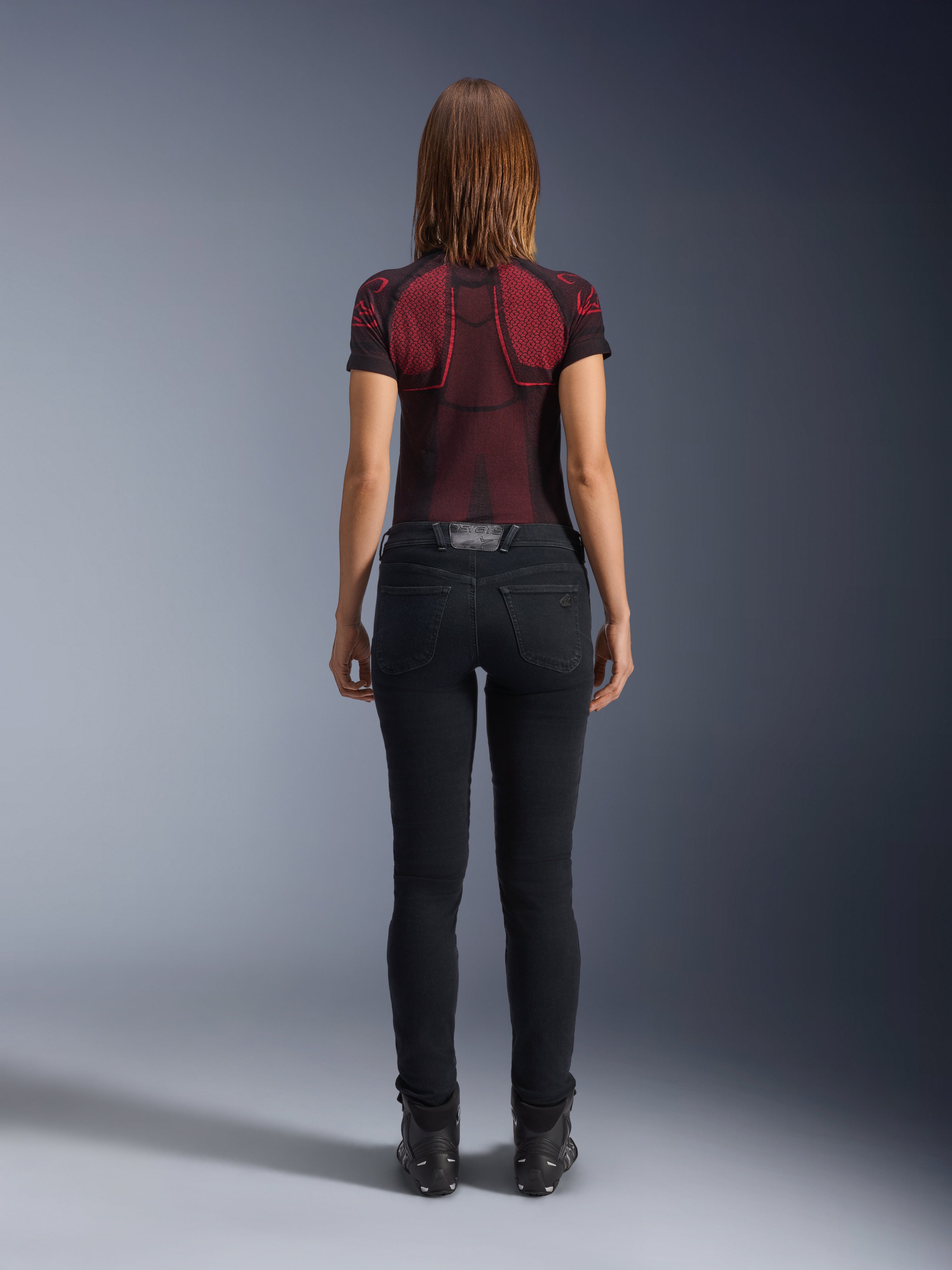 Alpinestars Daisy V3 dames motorjeans, Rinse Black, slim-fit motorbroek van achteren gefotografeerd, met een high-waist ontwerp en achterzakken, gecombineerd met een rood en zwart technisch shirt met patroon en zwarte laarzen, urban motorkleding voor dames