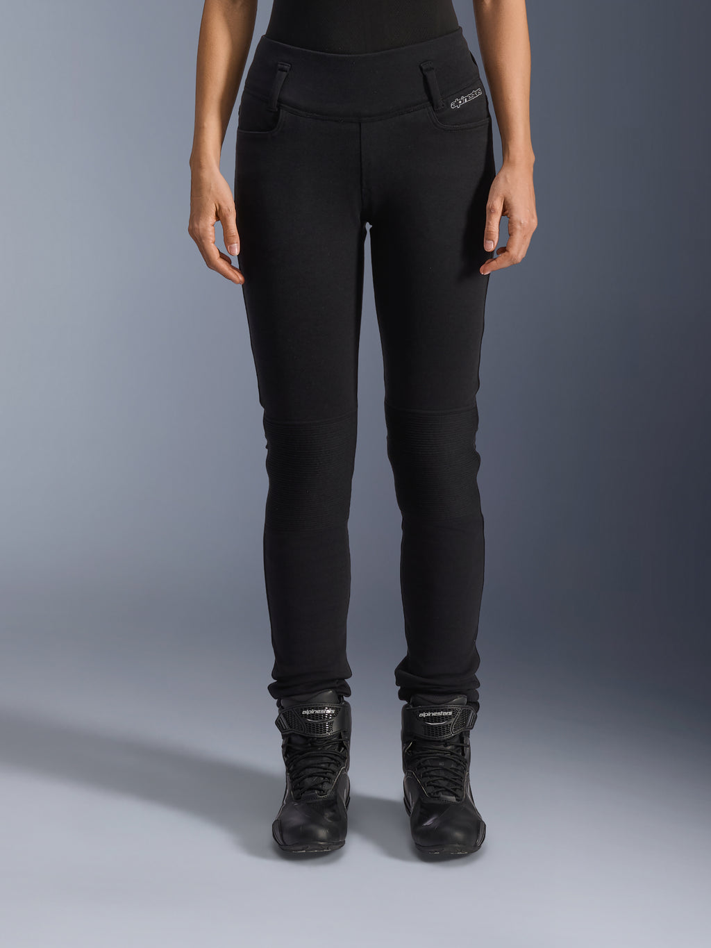 Alpinestars Womens Banshee Leggings, zwarte motorbroek voor dames, slim-fit ontwerp met accordeon-kniepanelen, gedragen met zwarte hoge rijlaarzen, urban motorkleding met een casual stijl