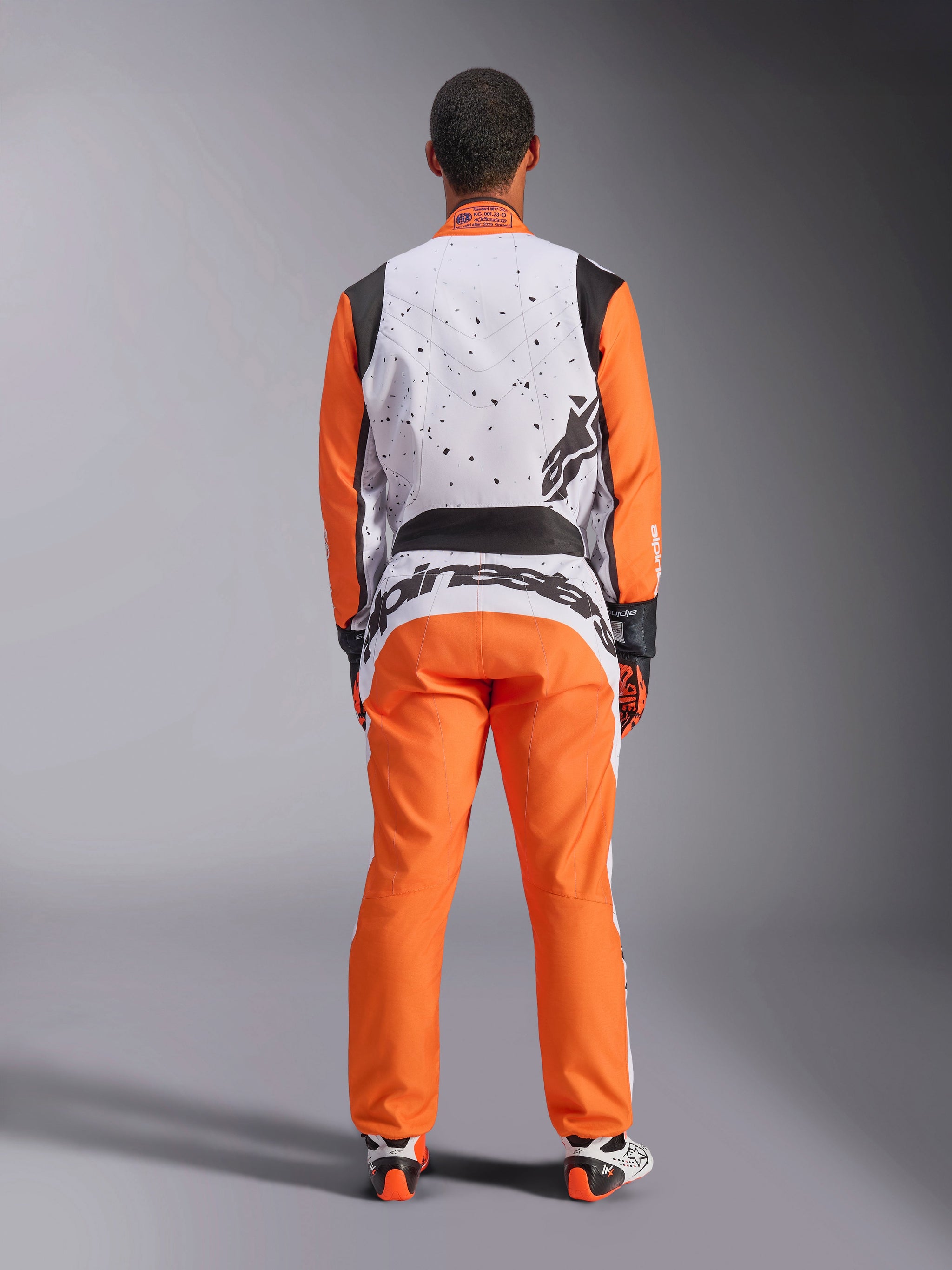 Rear view of a racer wearing the Alpinestars KMX-9 V3 Graphic 6 Suit in White Orange Black, white upper back with black speckle graphics, orange sleeves and pants, large black Alpinestars branding on the lower back, coordinated with racing gloves and boots for karting.