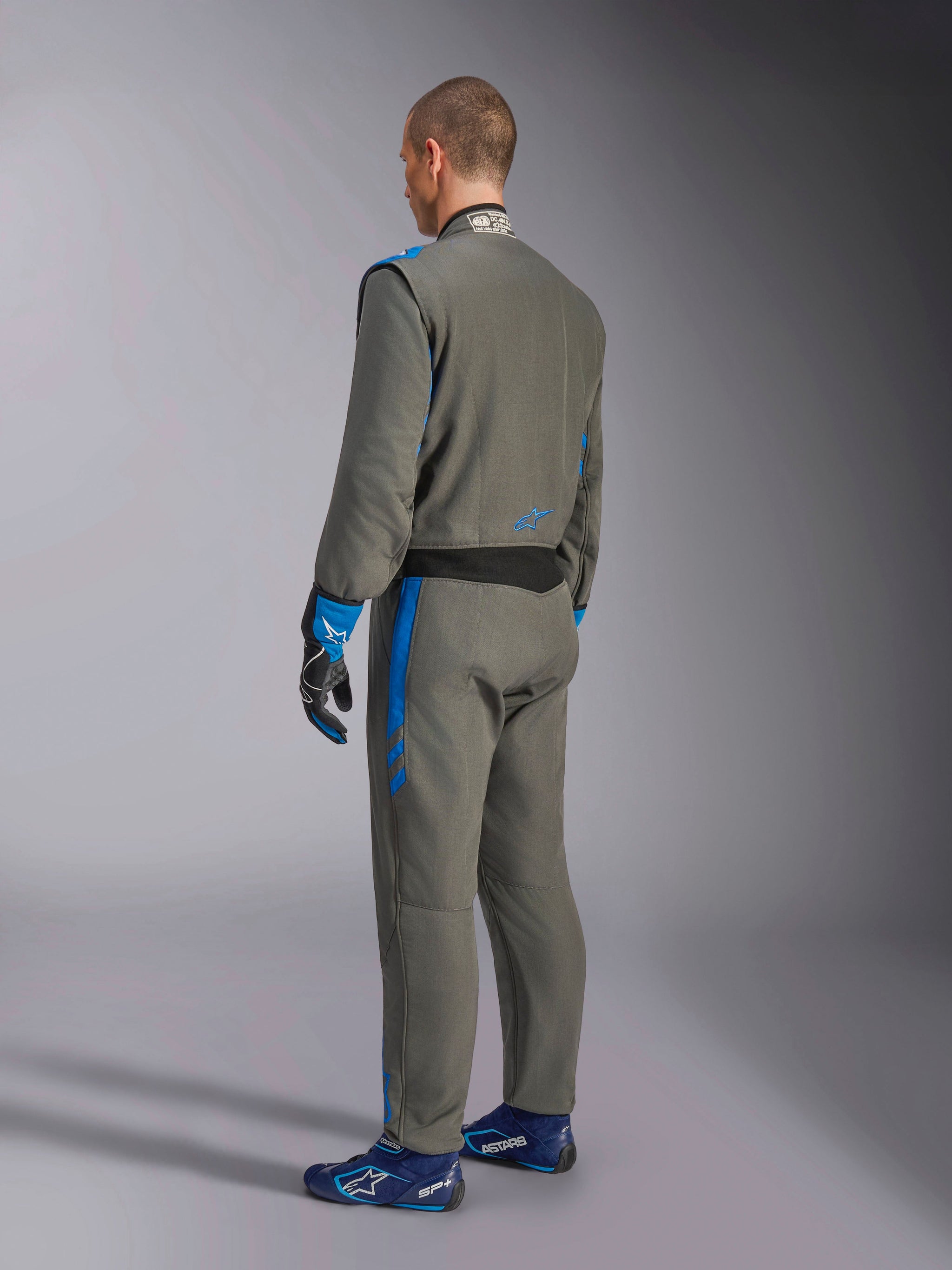 Alpinestars GP Race V3 Suit FIA, autoracepak, Antraciet blauw, achteraanzicht van model in het grijze eendelige pak met blauwe accenten, bijpassende blauwe handschoenen en raceschoenen, professioneel design voor motorsportuitrusting