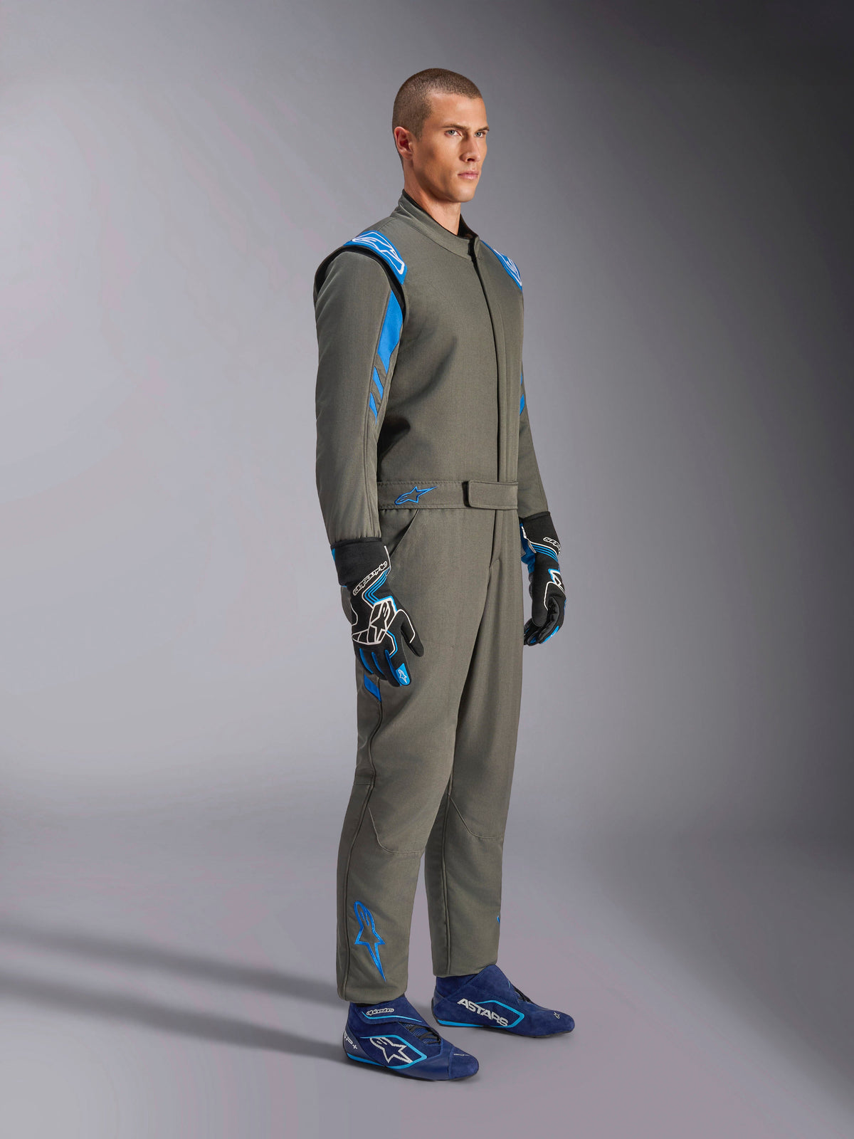 Alpinestars GP Race V3 Pak FIA in antracietblauw, eendelig autoracepak gedragen door een mannelijk model, voorzien van helderblauwe accenten op de schouders en enkels, gecombineerd met bijpassende racehandschoenen en blauwe raceschoenen, professionele autosportuitrusting.