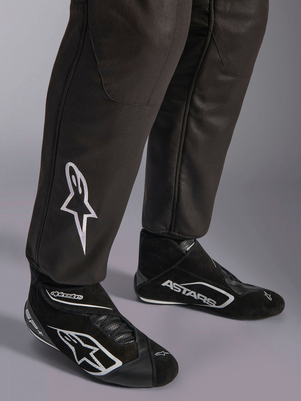 Alpinestars GP Race V3 Suit FIA, close-up of lower legs and racing boots, Black with Silver accents, featuring white embroidered star logo on the shin and ribbed ankle cuffs, professional auto racing protective gear.