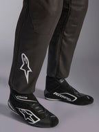 Alpinestars GP Race V3 Suit FIA, close-up of lower legs and racing boots, Black with Silver accents, featuring white embroidered star logo on the shin and ribbed ankle cuffs, professional auto racing protective gear.