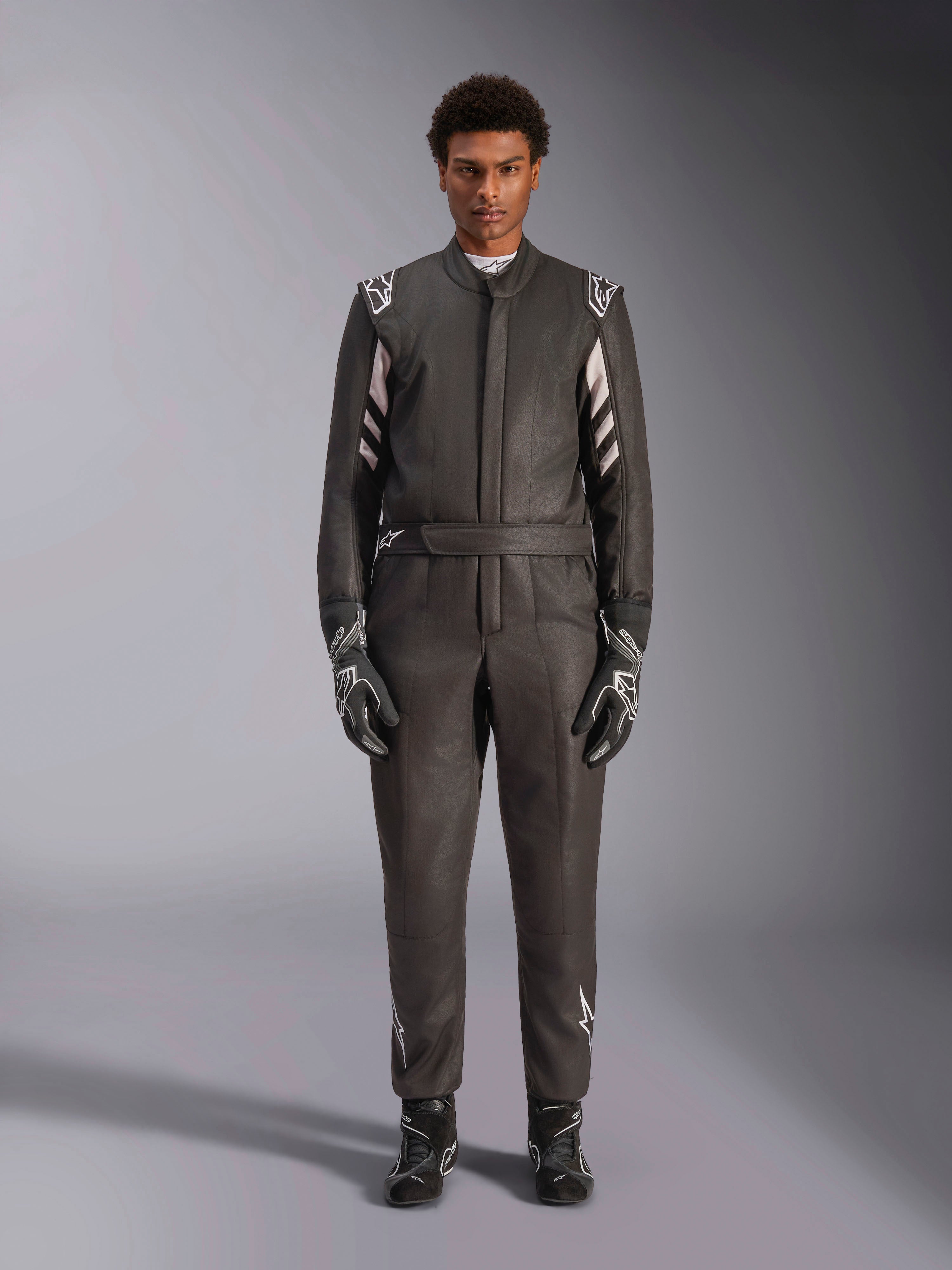 Alpinestars GP Race V3 Pak FIA, autoracepak, zwart en zilver, vooraanzicht van een coureur in volledige race-uitrusting inclusief handschoenen en laarzen, professioneel motorsportontwerp met Alpinestars-branding op de schouders en riem