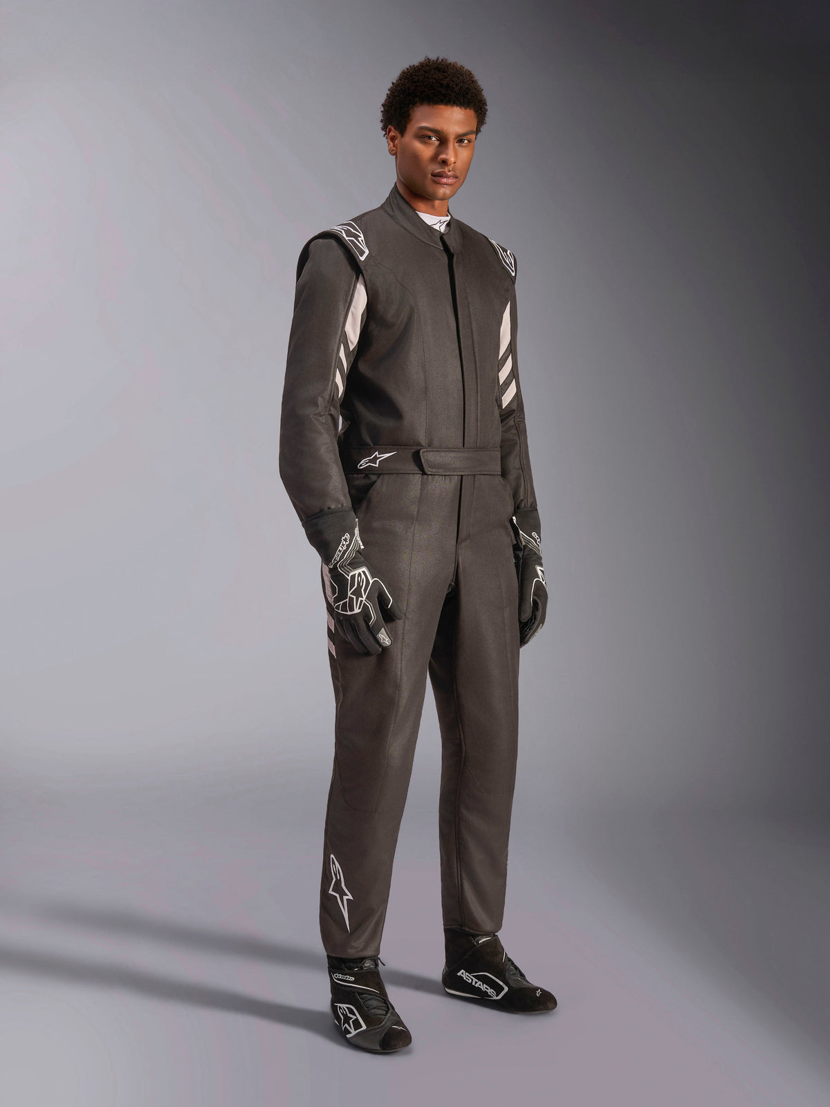 Mannelijke coureur in Alpinestars GP Race V3 Suit FIA, zwart en zilveren autoracepak, compleet met bijpassende handschoenen en schoenen, voorzien van zilveren accenten op de schouders en het onderbeen, professionele autorace-uitrusting met een getailleerde pasvorm.