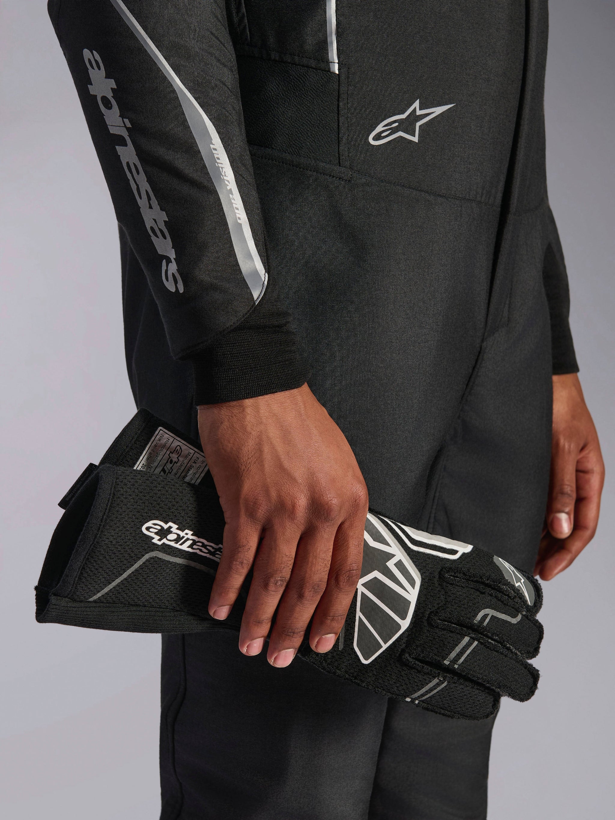 Alpinestars Hypertech V3-pak in Black Dark Shadow, close-up detail van de mouw en torso met zilveren Alpinestars-branding, hand die een zwarte racehandschoen met witte accenten vasthoudt, professionele autorace-uitrusting