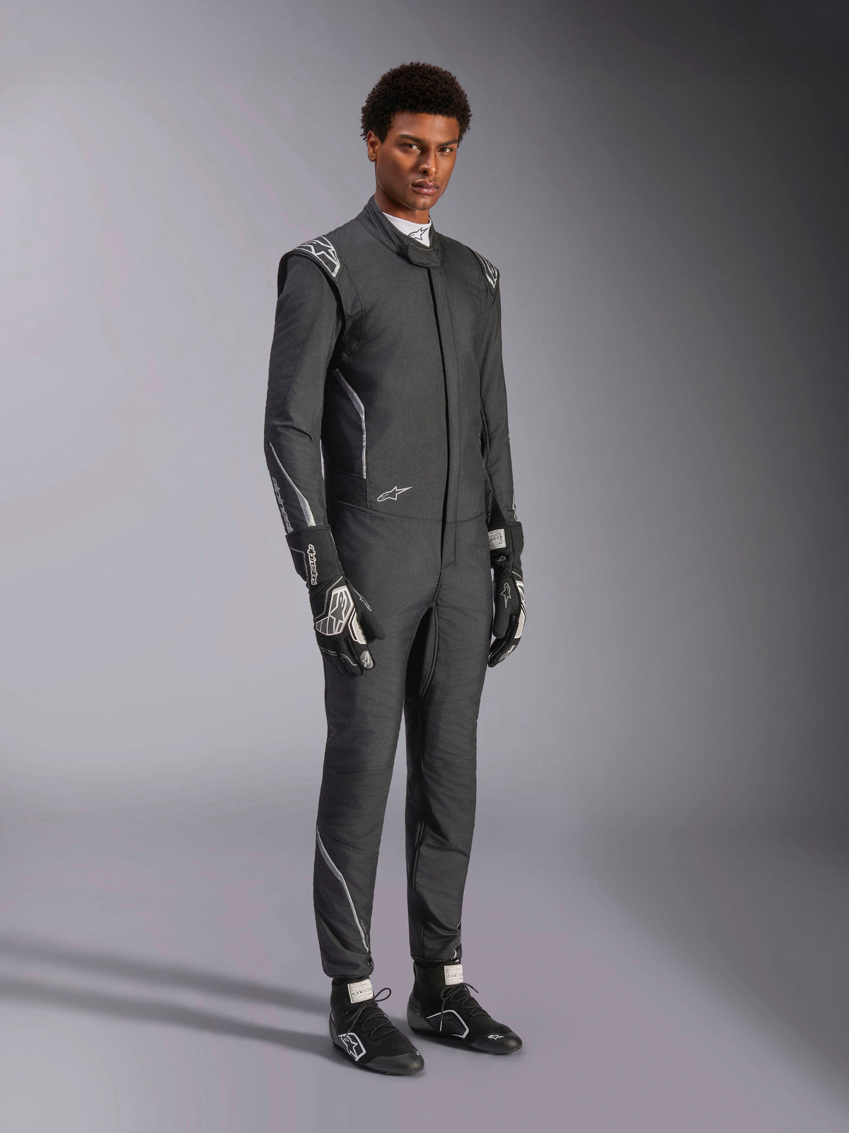Alpinestars Hypertech V3-pak, autoracepak, zwart en Dark Shadow, gedragen door mannelijk model met bijpassende zwarte handschoenen en raceschoenen, atletische slanke pasvorm, voorzien van Alpinestars-logo's en lichtgrijze accenten, professionele beschermende motorsportuitrusting