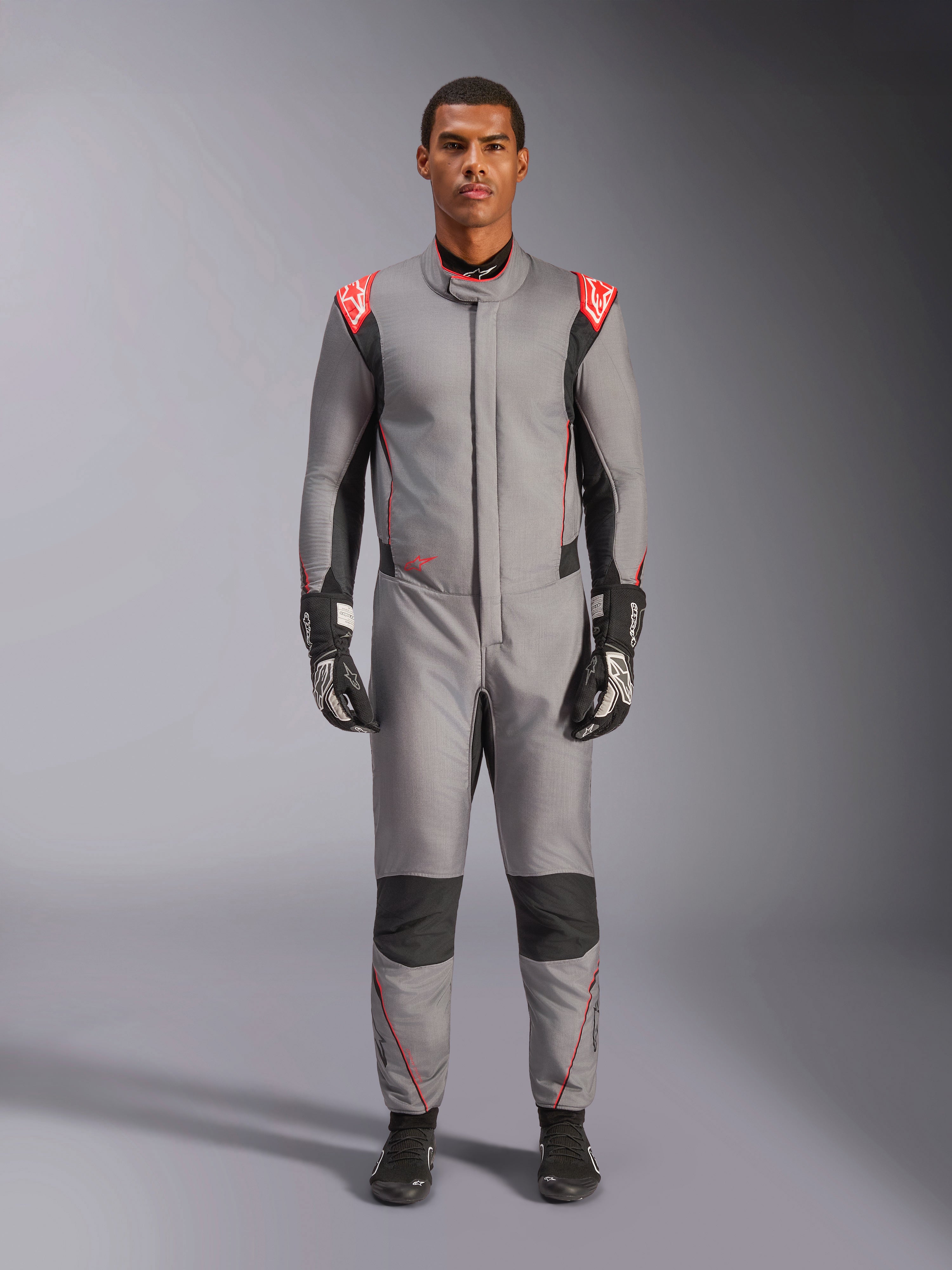 Alpinestars Hypertech V3 Suit, professioneel autoracepak in Mid Gray Black en Red Fluo, gedragen door een model met zwarte racehandschoenen en schoenen, atletische slanke pasvorm, fluo rode schouderaccenten, zwarte panelen op de knieën en zijkanten.