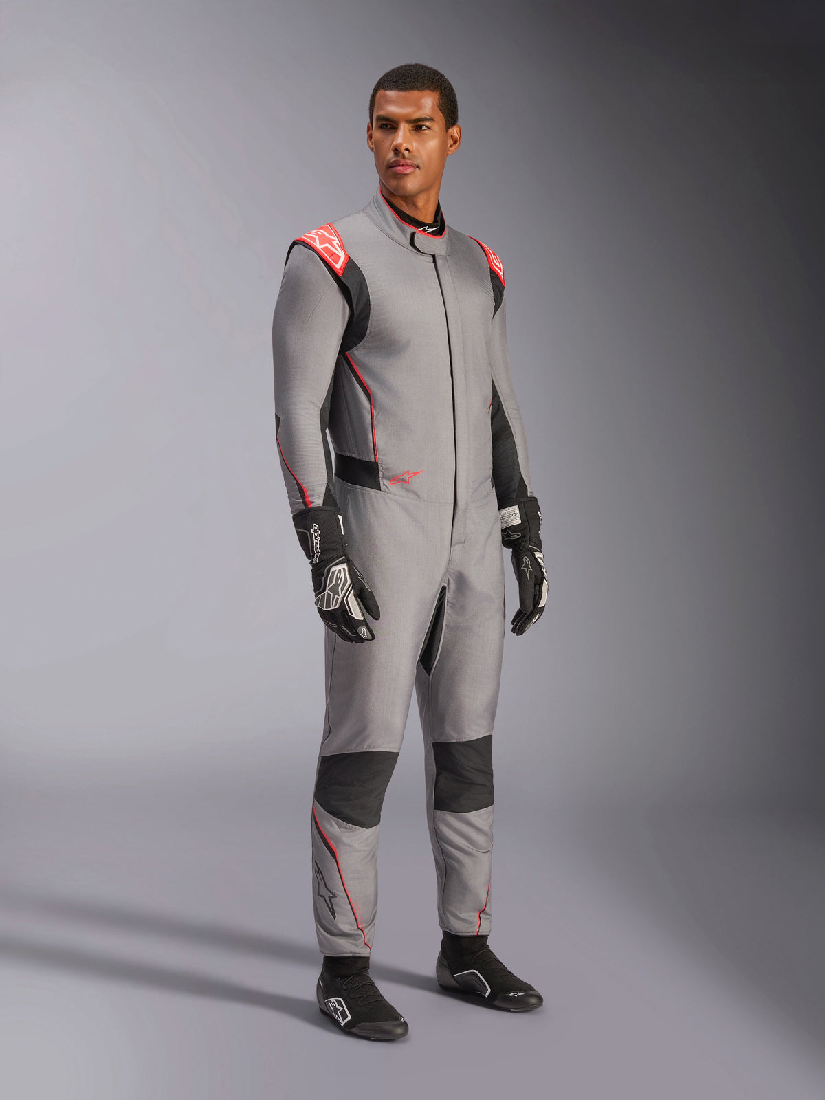 Mannelijk model draagt het Alpinestars Hypertech V3-pak in Mid Gray met zwarte en Red Fluo accenten, professioneel autoracepak gecombineerd met zwarte racehandschoenen en schoenen, voorzien van een lichtgewicht tweelaagse Nomex-constructie met rode schouderdetails en een atletische pasvorm.