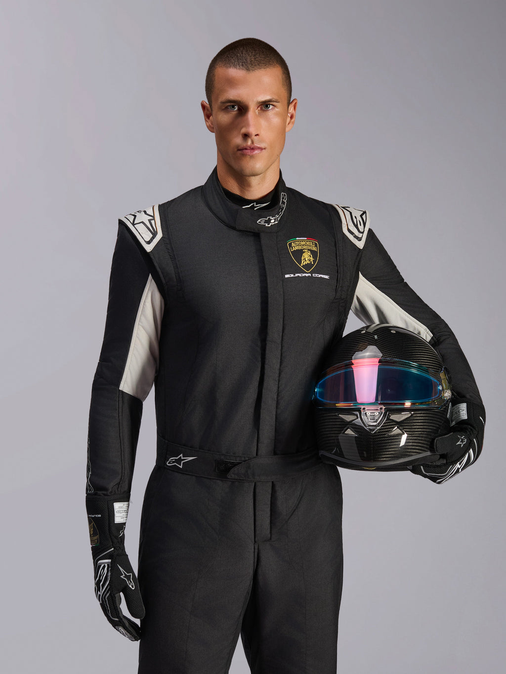 Male model wearing Limited Edition Lamborghini GP Tech V4 Suit, black racing suit with silver accents and Lamborghini Squadra Corse logo, holding a carbon fiber helmet, black racing gloves with white logos, professional auto racing gear