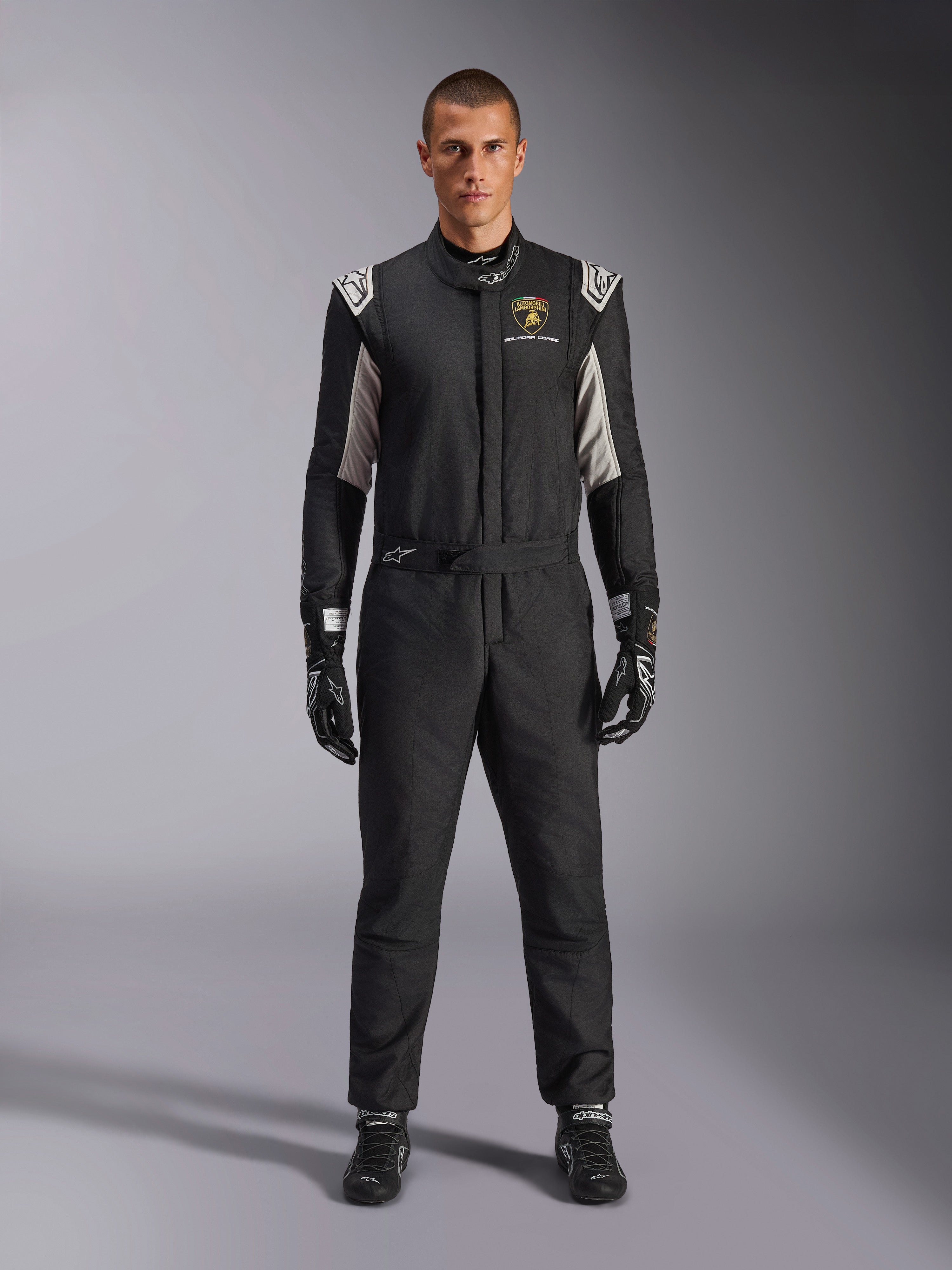 Alpinestars Limited Edition Lamborghini GP Tech V4 Pak FIA, professioneel autoracepak, zwart met zilveren en gouden accenten, voorzien van Automobili Lamborghini Squadra Corse-logo op de borst, gedragen met bijpassende racehandschoenen en laarzen, strak atletisch ontwerp voor circuitwedstrijden