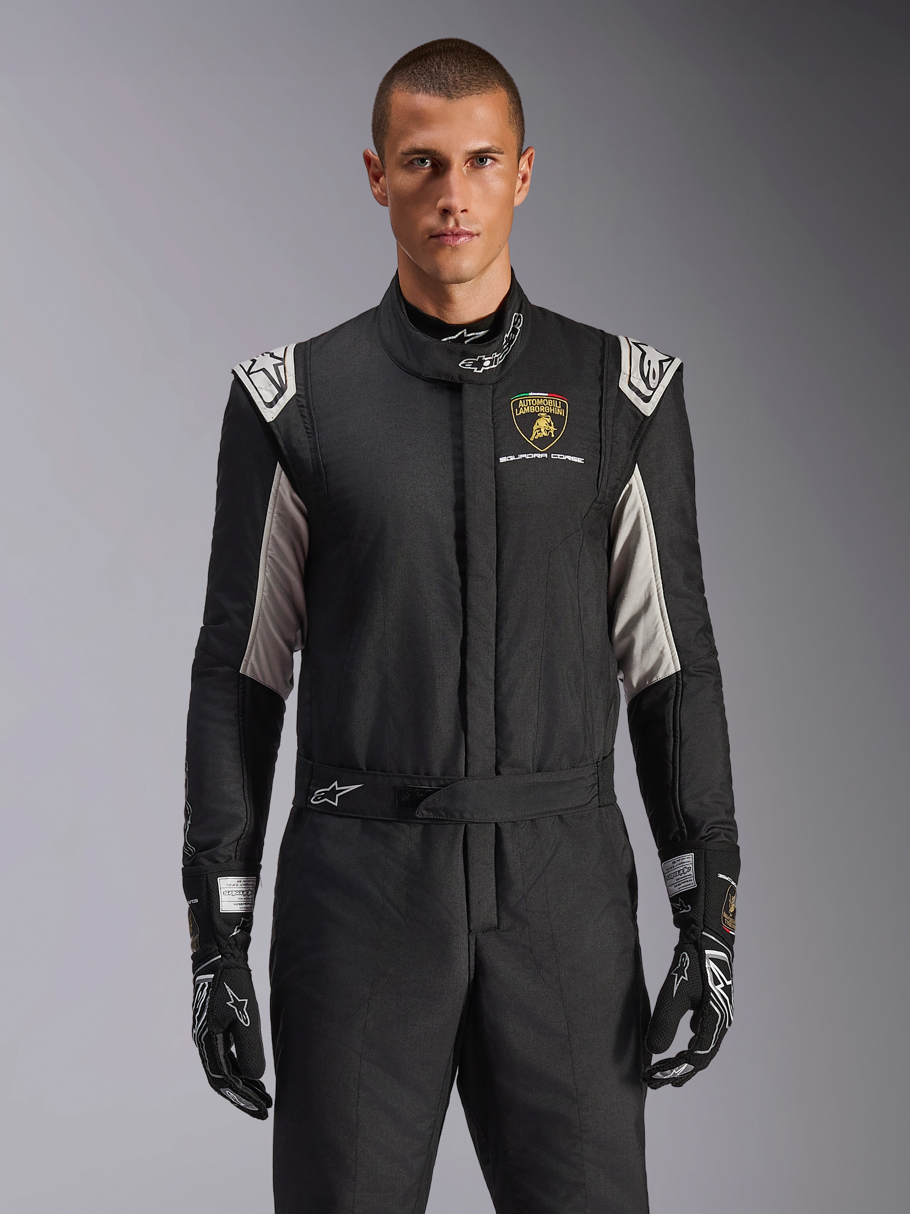 Alpinestars Limited Edition Lamborghini GP Tech V4 Pak, professioneel racepak, zwart met zilveren en gouden accenten, voorzien van het Automobili Lamborghini Squadra Corse-schild op de borst, gedragen door een model met bijpassende zwarte racehandschoenen