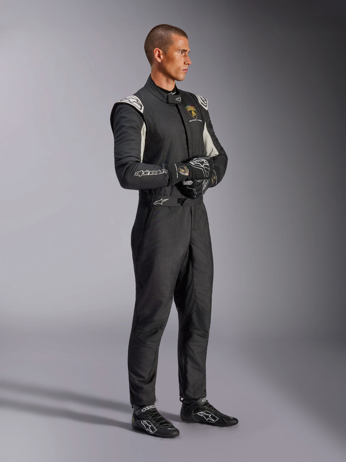 Mannelijke coureur in het Alpinestars Limited Edition Lamborghini GP Tech V4-pak, FIA-gecertificeerd autoracpak in zwart, zilver en goud, gecombineerd met bijpassende racehandschoenen en schoenen, voorzien van Lamborghini Squadra Corse-branding, professionele motorsportuitrusting