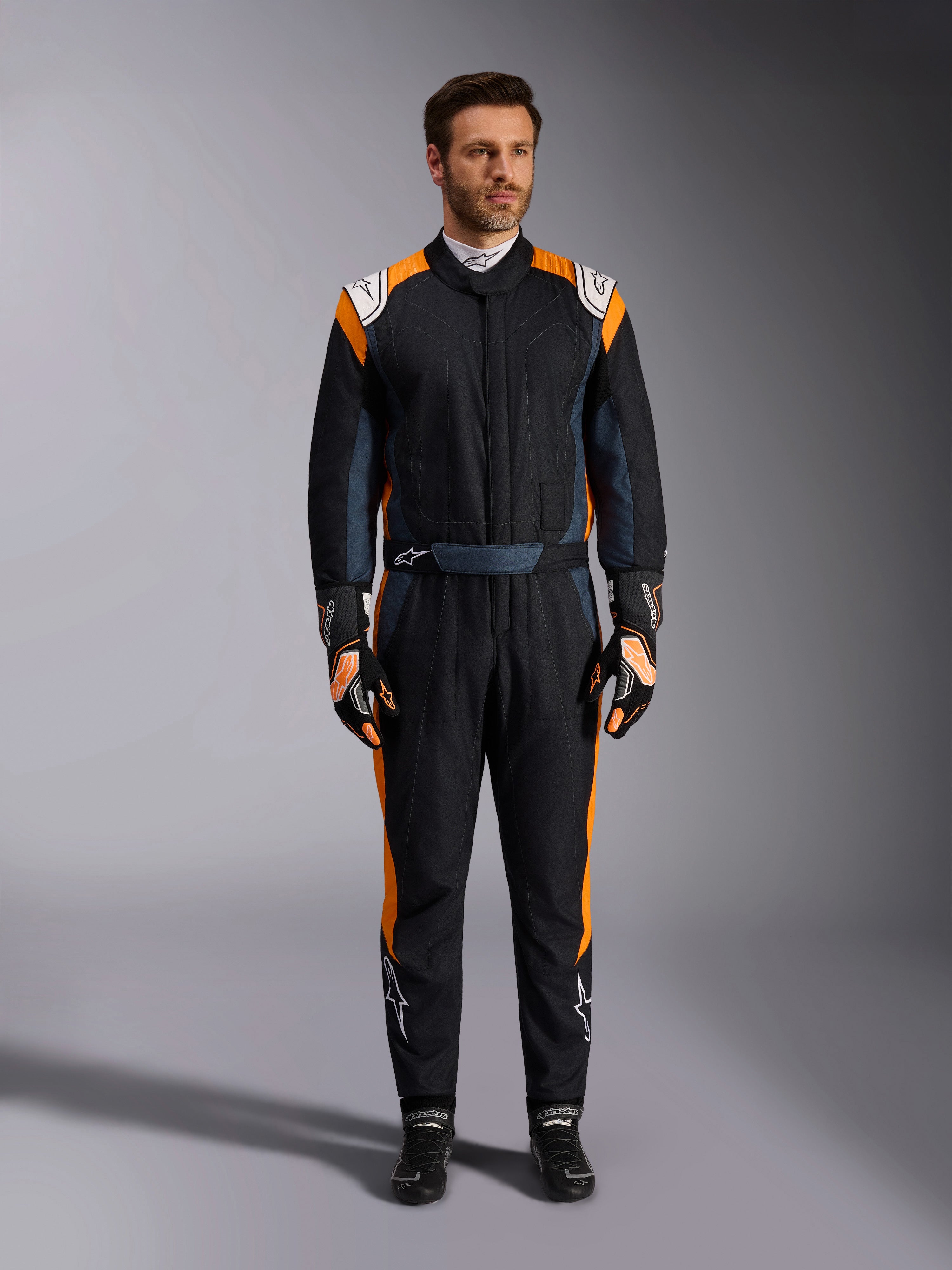 Alpinestars GP Pro Comp V2 FIA Suit, Black Asphalt en Orange Fluo autoracepak, mannelijk model met volledige beschermende uitrusting, inclusief bijpassende racehandschoenen en -schoenen, voorzien van witte Alpinestars-logo's en opvallende oranje zijpanelen, professionele motorsportkleding