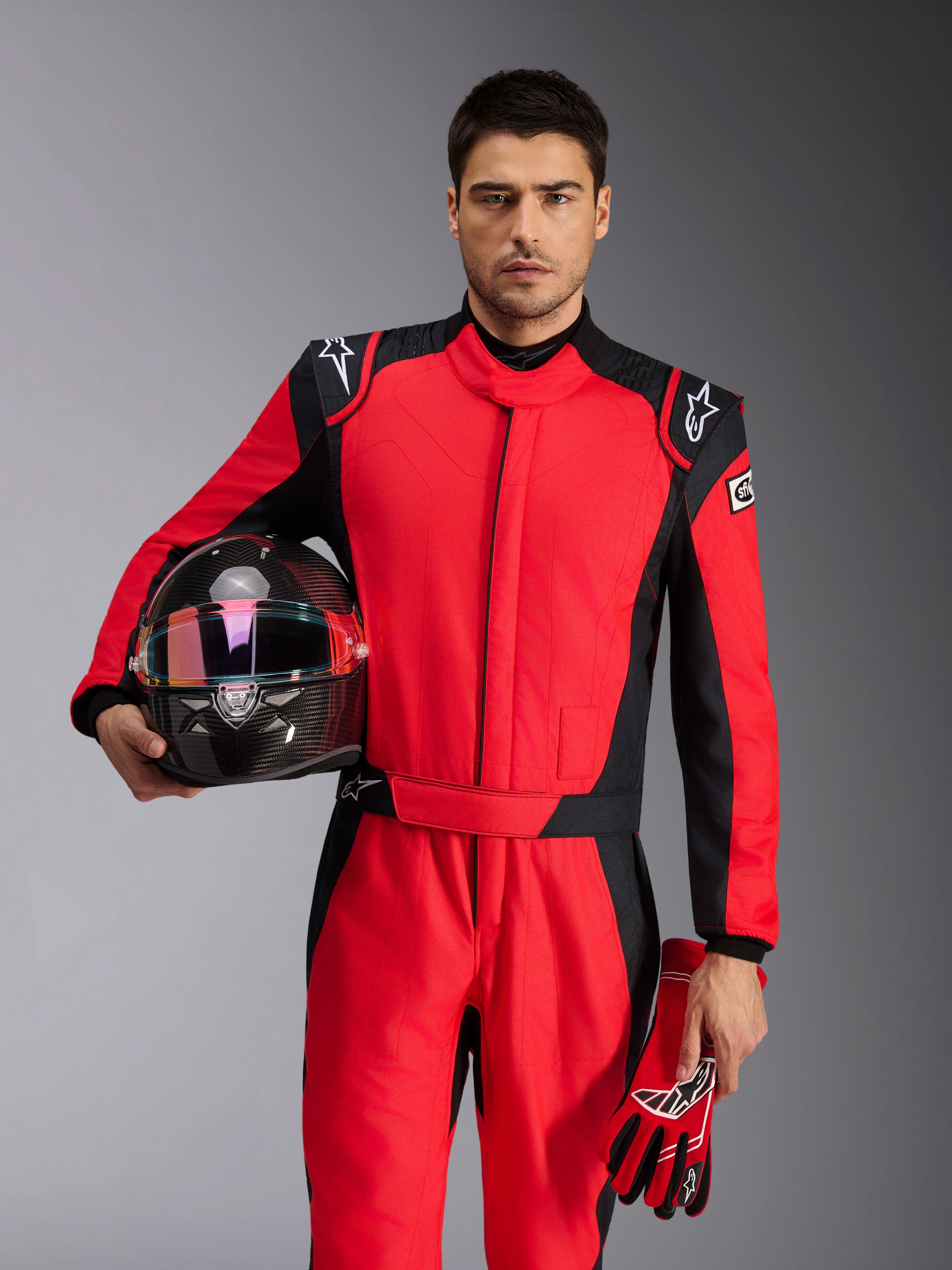 Mannelijke autocoureur in Alpinestars GP Pro Comp V2 FIA Pak in rood en zwart, houdt een koolstofvezel racehelm en bijpassende handschoenen vast, professionele autosportuitrusting