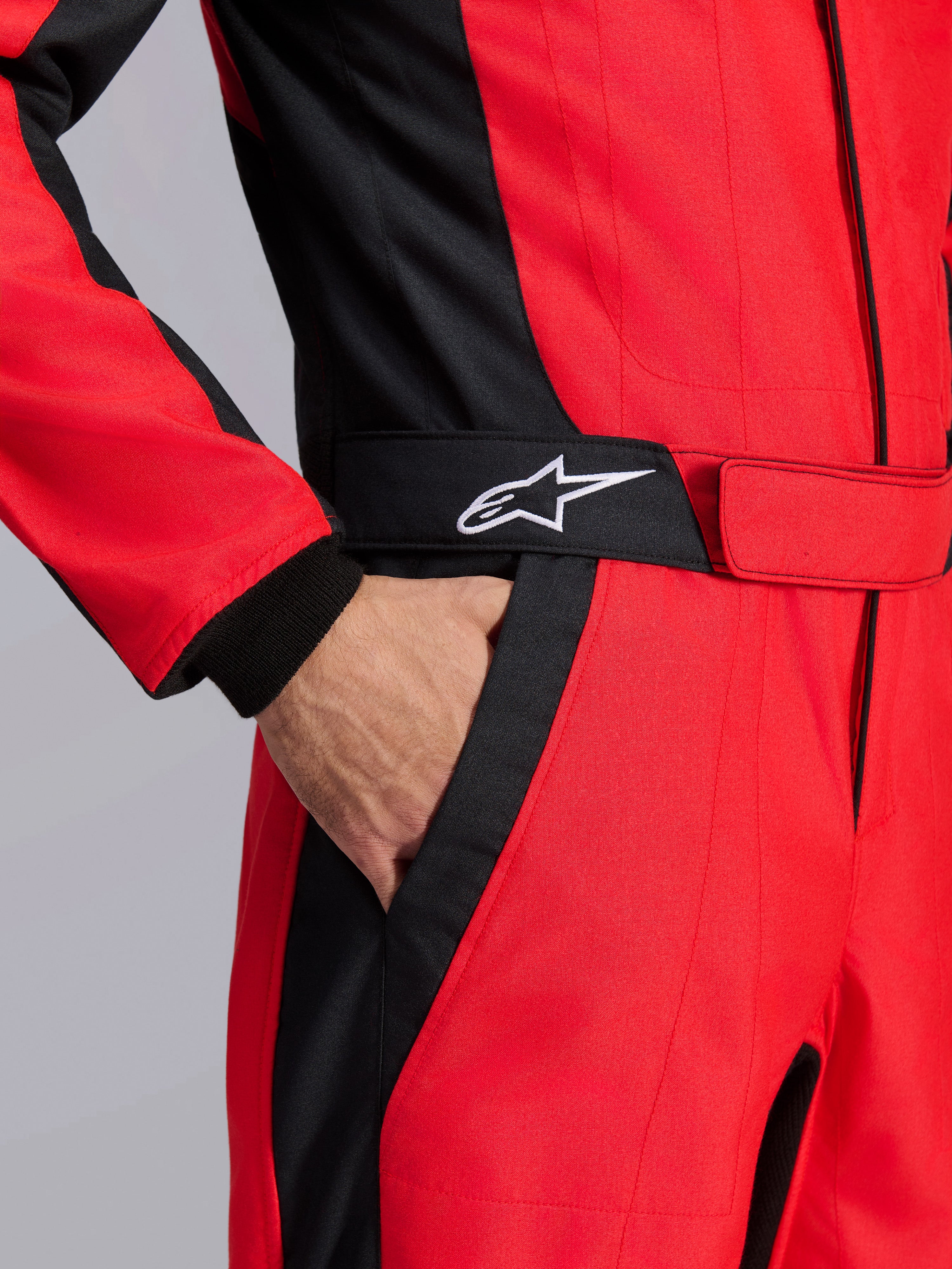 Alpinestars GP Pro Comp V2 FIA racepak, rood en zwart, close-up van de taille met een hand in de zak, zwarte riem met wit sterlogo en gestructureerde panelen voor autoracen