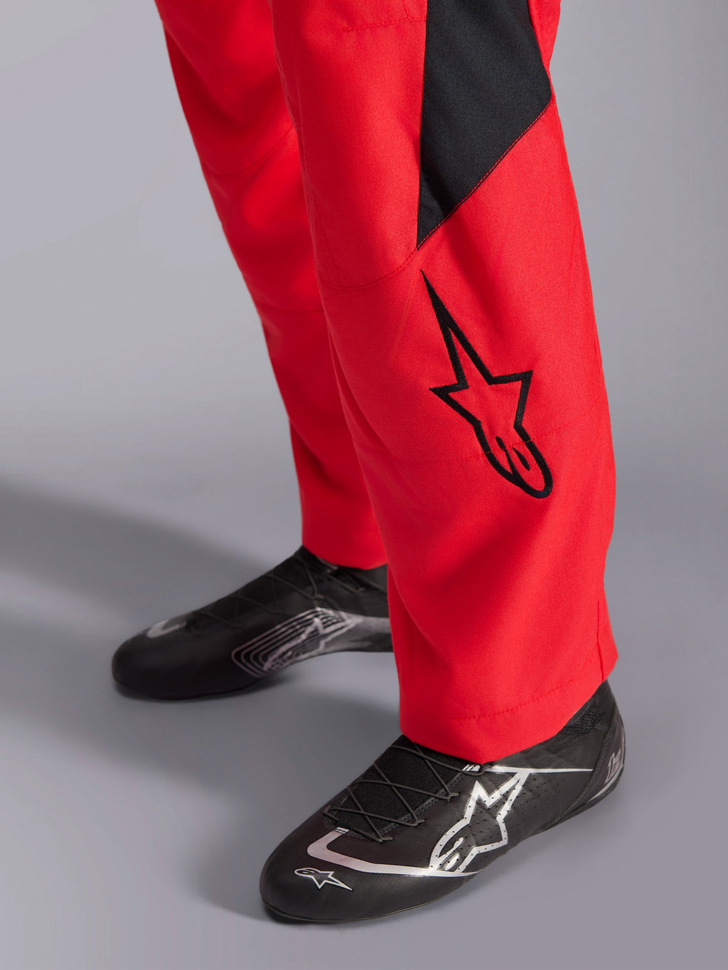 Alpinestars GP Pro Comp V2 FIA racepak, close-up van het onderbeen en de raceschoenen, rood met een zwart Astars-logo en paneel, gecombineerd met zwarte performance raceschoenen, autorace-uitrusting.
