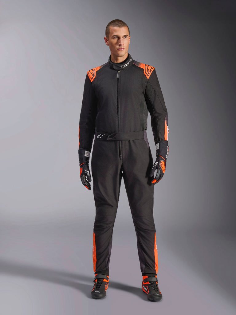 Alpinestars KMX-5 V3 Suit, zwart en fluo oranje kartpak, gedragen door een model met bijpassende racehandschoenen en schoenen, voorzien van oranje accenten op de schouders en benen, professioneel eendelig motorsportontwerp met een technische mesh-uitstraling