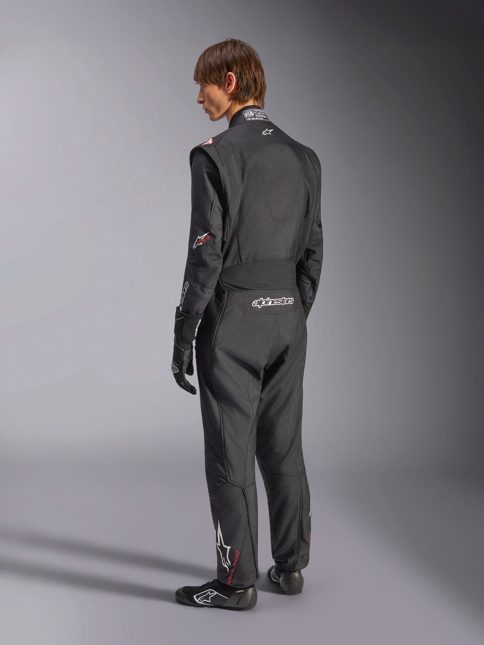Alpinestars GP Tech V4-pak, autoracepak, zwart met witte accenten, achteraanzicht van model dat het pak draagt met racehandschoenen en -schoenen, voorzien van witte branding op de onderrug en het been, professionele autosport veiligheidsuitrusting