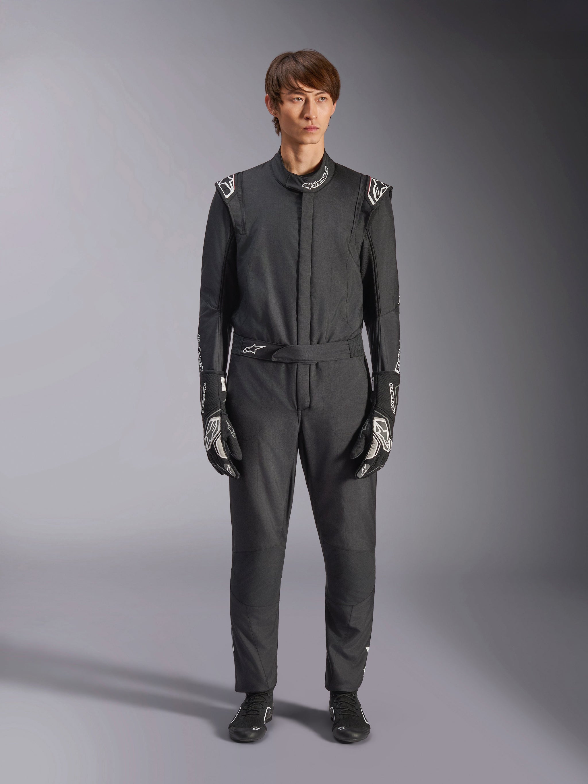 Alpinestars GP Tech V4 Suit, zwart en wit, professioneel eendelig autoracepak, gedragen met bijpassende zwart-witte handschoenen en raceschoenen, met witte Alpinestars-logo's op de schouders en kraag, strakke atletische pasvorm voor motorsportwedstrijden