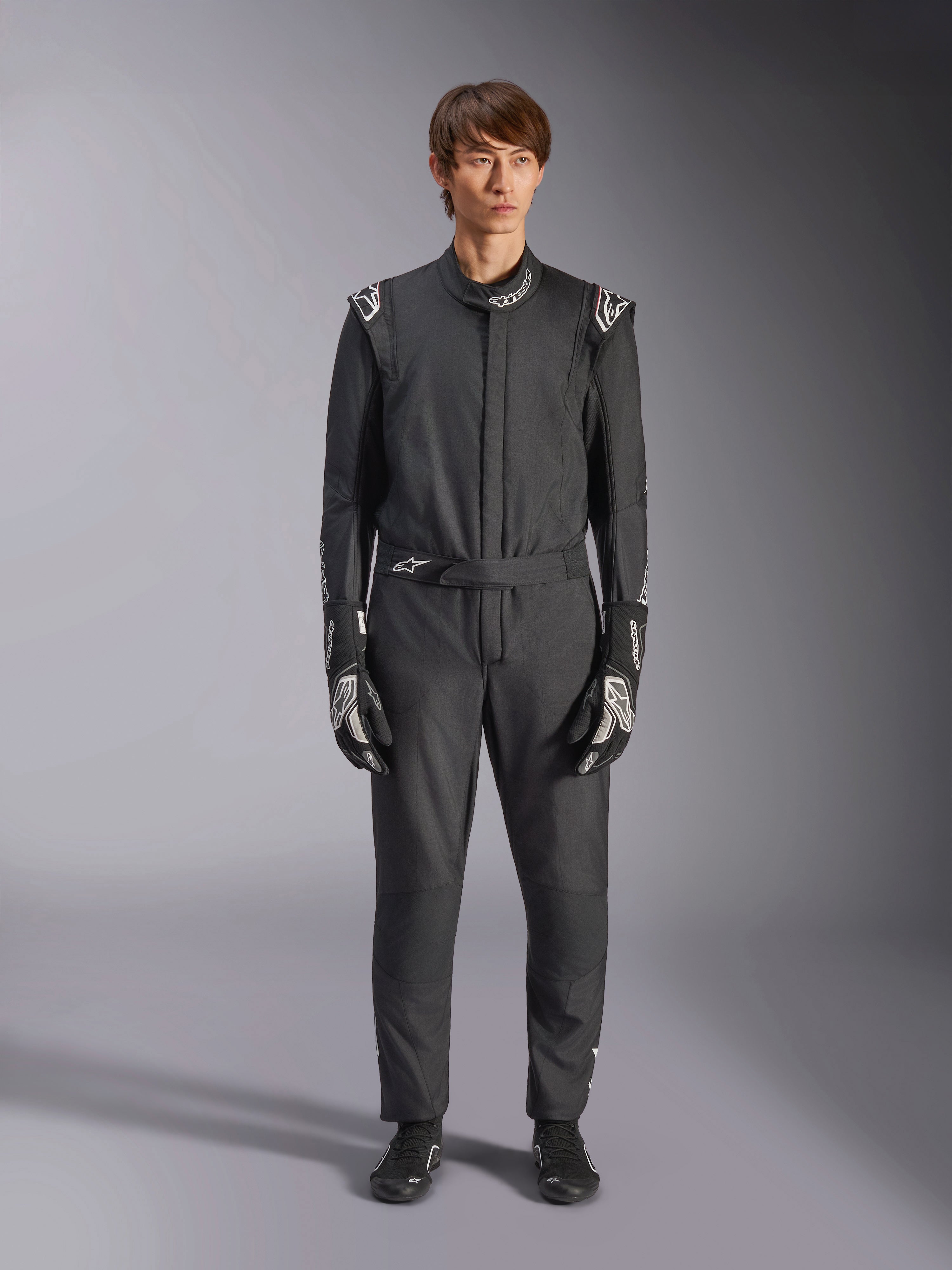 Alpinestars GP Tech V4 Suit, zwart en wit, professioneel eendelig autoracepak, gedragen met bijpassende zwart-witte handschoenen en raceschoenen, met witte Alpinestars-logo's op de schouders en kraag, strakke atletische pasvorm voor motorsportwedstrijden