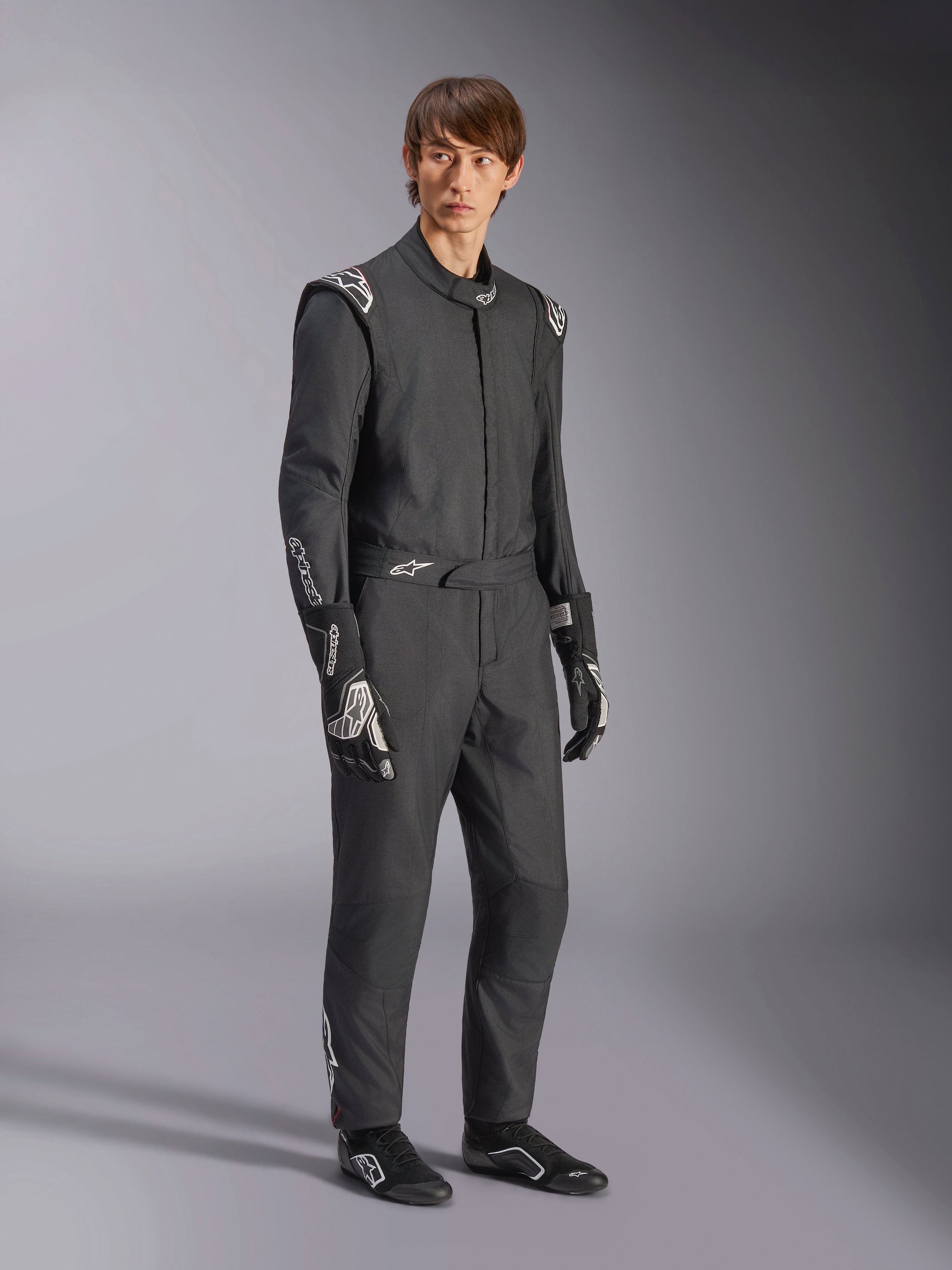 Alpinestars GP Tech V4 Suit, eendelig autoracepak, zwart en wit, gedragen met bijpassende racehandschoenen en schoeisel, voorzien van witte logo's op de schouders en onderarmen, strak professioneel ontwerp voor autosport