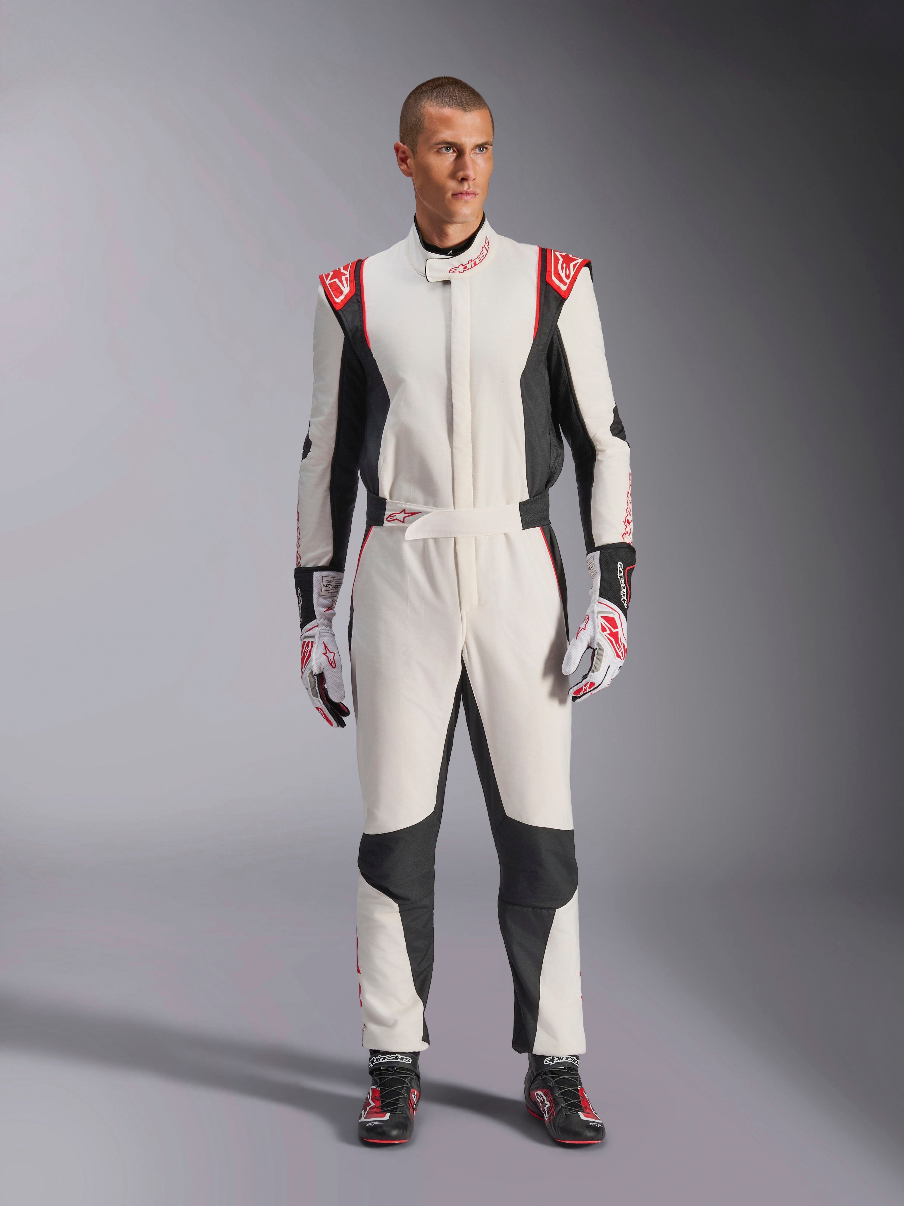 Alpinestars GP Tech V4 Suit, wit zwart en rood eendelig autoracepak, gedragen door een mannelijk model met bijpassende racehandschoenen en -schoenen, met rode logo's op de schouders en zwarte zijpanelen, professionele motorsportuitrusting voor circuitgebruik