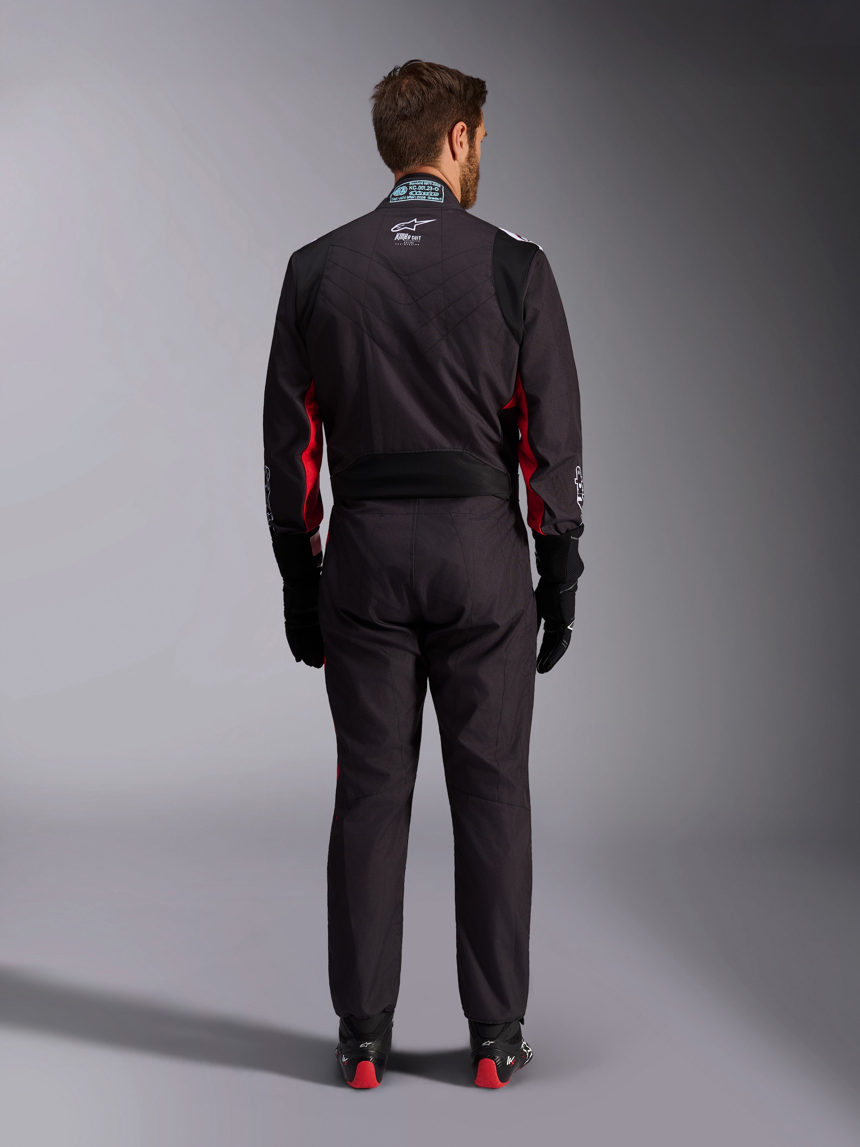 Alpinestars KMX-9 V3 Graphic 3 pak, karting pak, zwart en rood, achteraanzicht met het lumbale stretchpaneel en FIA-certificeringslabel op de kraag, gestyled met zwarte racehandschoenen en rood geaccentueerde laarzen
