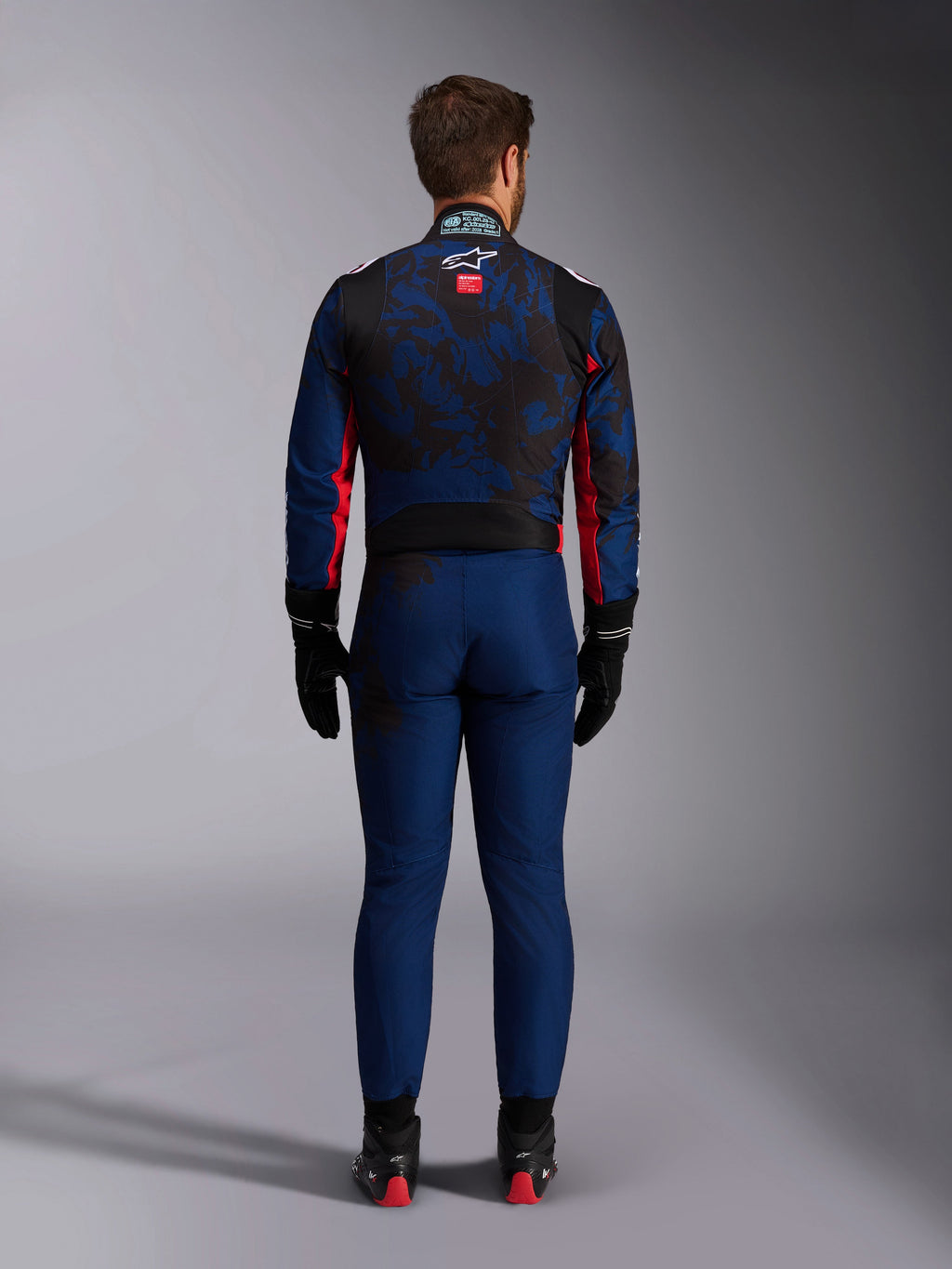 Kartingcoureur van achteren gezien in het KMX-9 V3 Graphic 5-pak in Navy Red, met een donkerblauw en zwart grafisch patroon, rode zij-accenten en het Alpinestars-logo op de bovenrug, gecombineerd met zwarte racehandschoenen en schoeisel.