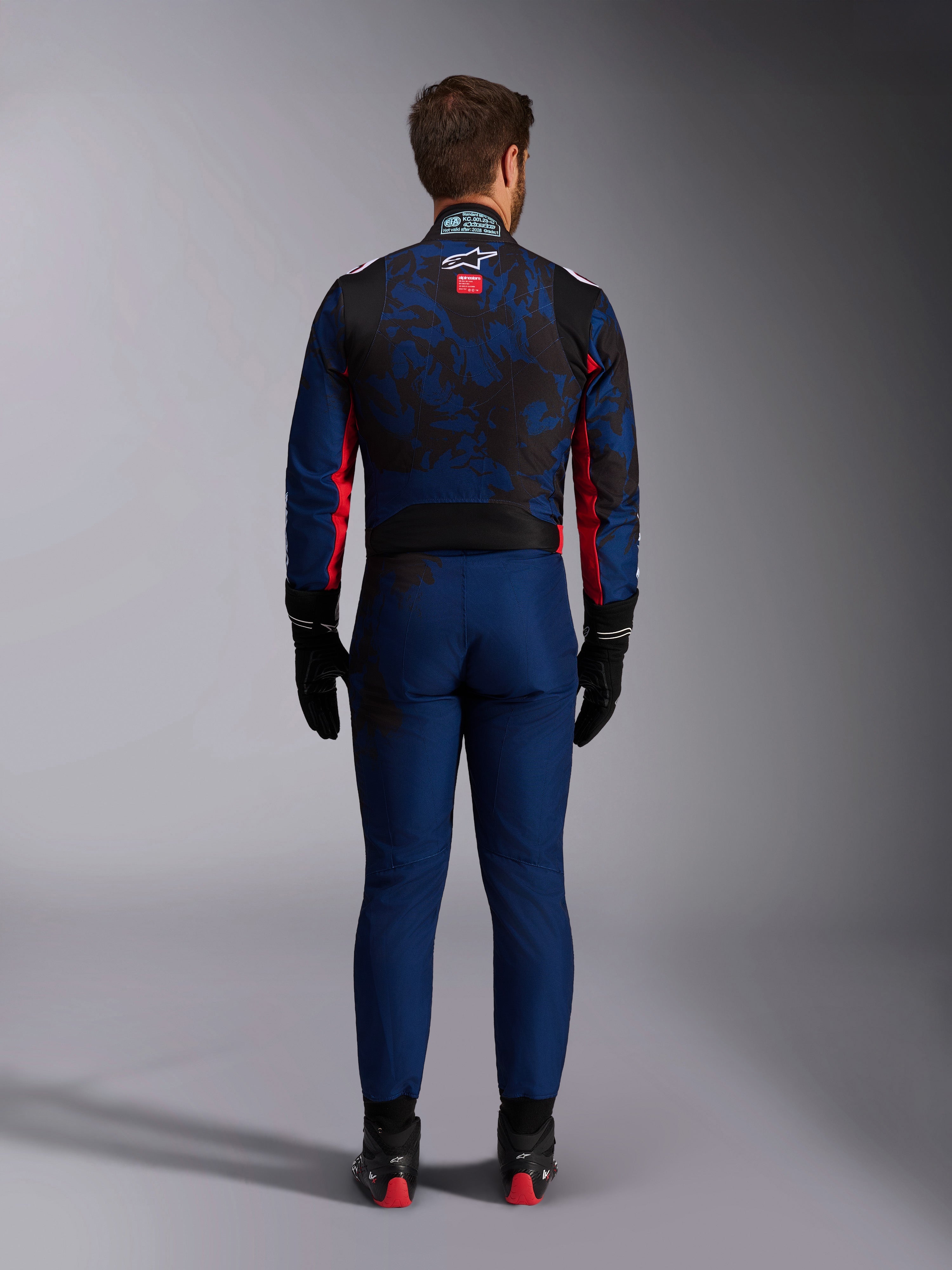Kartingcoureur van achteren gezien in het KMX-9 V3 Graphic 5-pak in Navy Red, met een donkerblauw en zwart grafisch patroon, rode zij-accenten en het Alpinestars-logo op de bovenrug, gecombineerd met zwarte racehandschoenen en schoeisel.