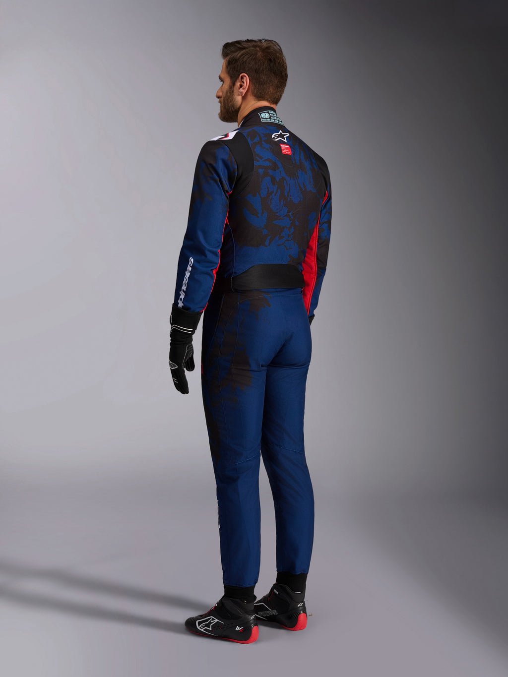 Alpinestars KMX-9 V3 Graphic 5 Suit, kartpak, Navy Rood, achteraanzicht met een marineblauwe basis, een donkere op camouflage geïnspireerde graphic op de rug en rode accenten, gedragen met zwarte racehandschoenen en zwart-rode kartingschoenen.