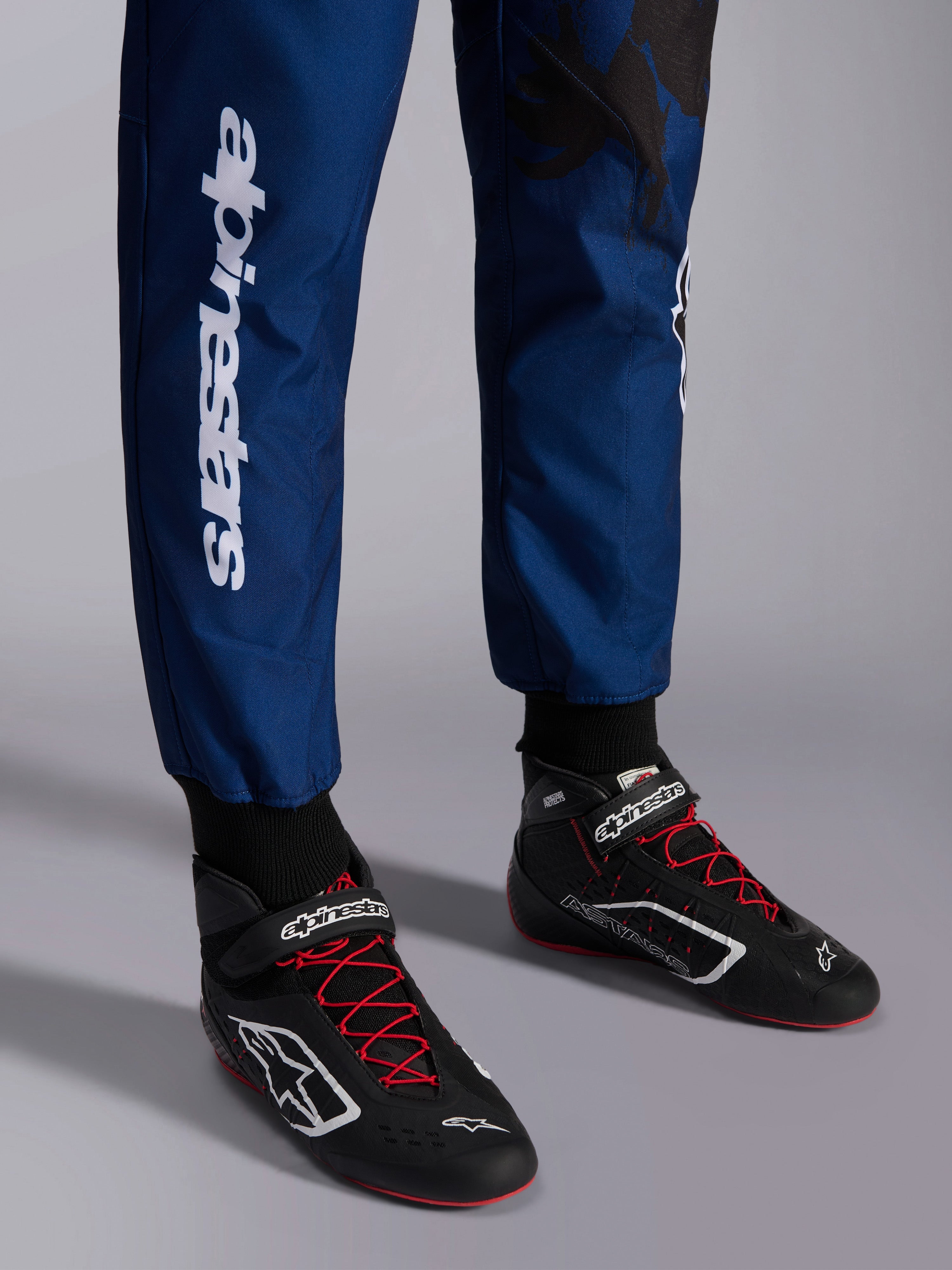 Alpinestars KMX-9 V3 Graphic 5 pak, Navy Rood, close-up van de onderbenen en kartschoenen, marineblauwe stof met witte verticale Alpinestars-branding, zwarte schoenen met rode veters en witte logo's, professionele kartinguitrusting