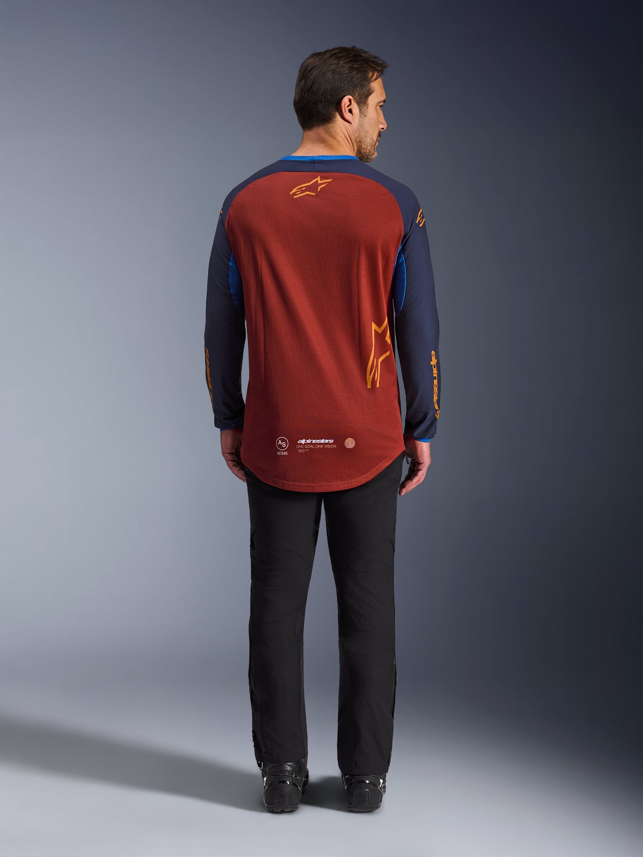 Alpinestars Maxdura Dual Jersey in Rust Brown and Mood Indigo, back view showing the dropped rear hem and logo detailing, worn with black technical pants and boots, designed for adventure and dual-sport motorcycling.