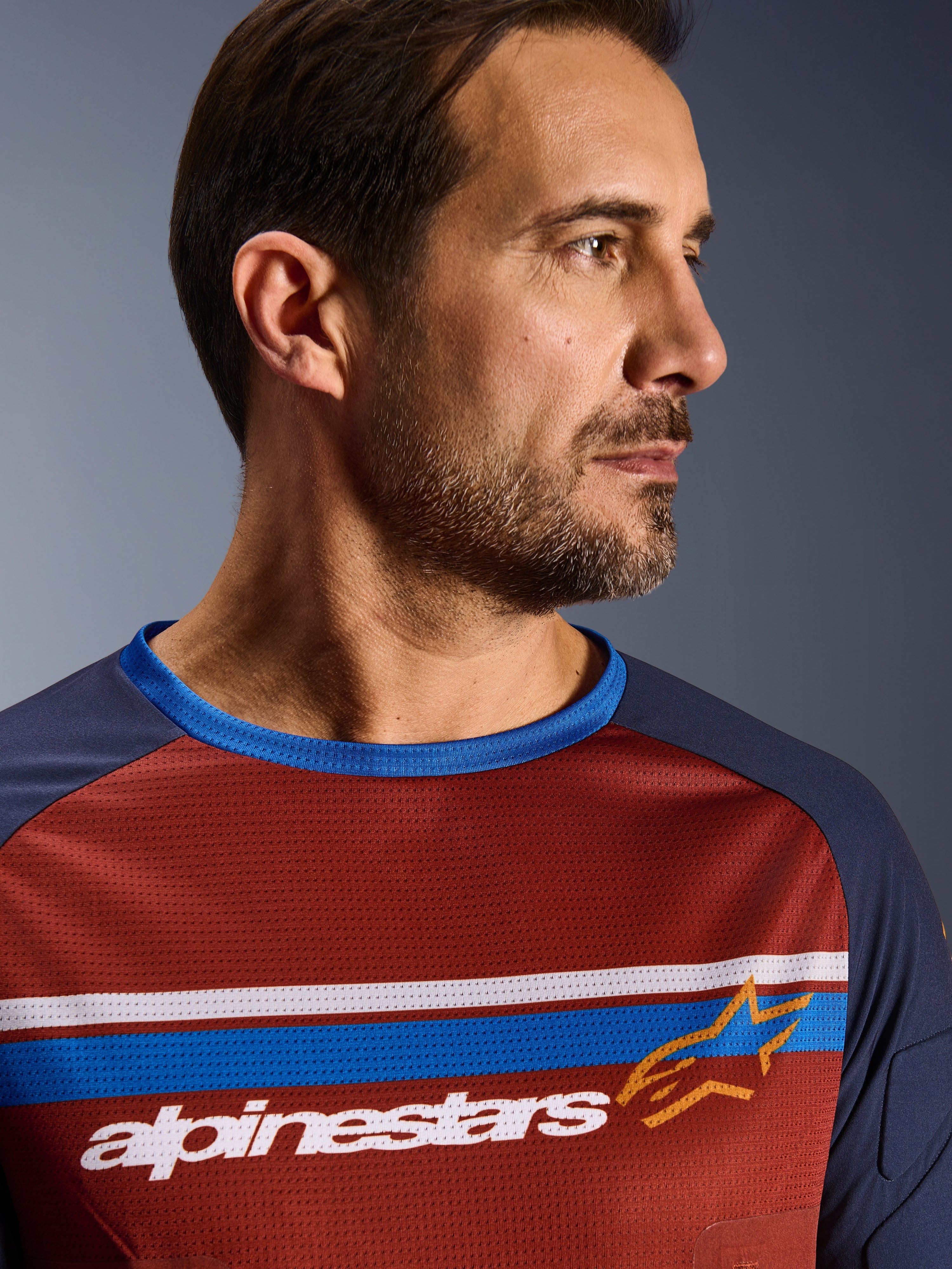 Alpinestars Maxdura Dual Jersey, Rustbruin en Mood Indigo, close-up van een rijder die de technische mesh jersey draagt, voorzien van horizontale witte en blauwe borststrepen, witte Alpinestars-branding en een oranje ster-logo.