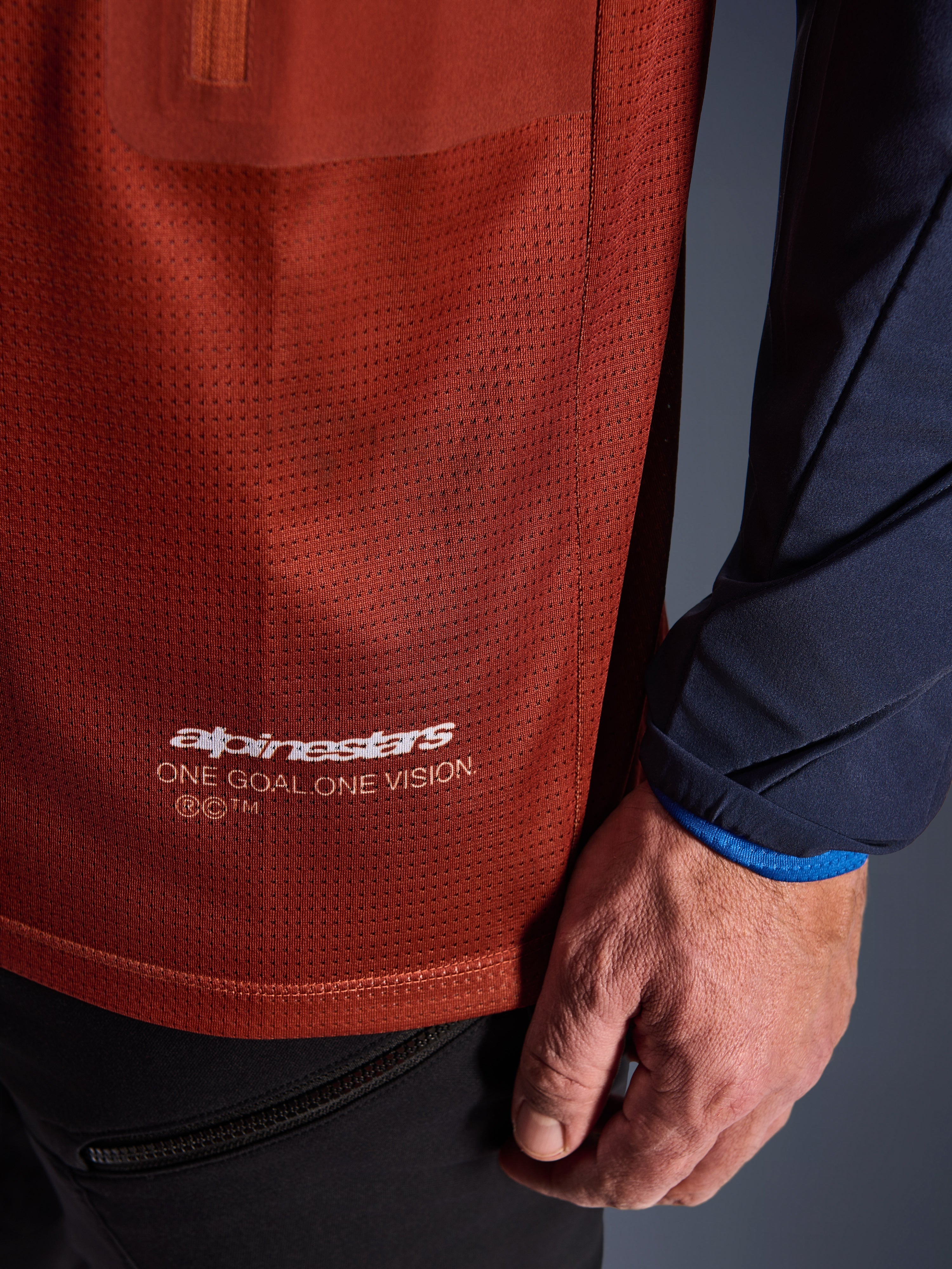 Alpinestars Maxdura Dual Jersey, adventure motorjersey, roestbruin en Mood Indigo, close-up van de mesh torso-stof en donkerblauwe geweven mouw, witte Alpinestars 'One Goal One Vision' branding op de onderste zoom.