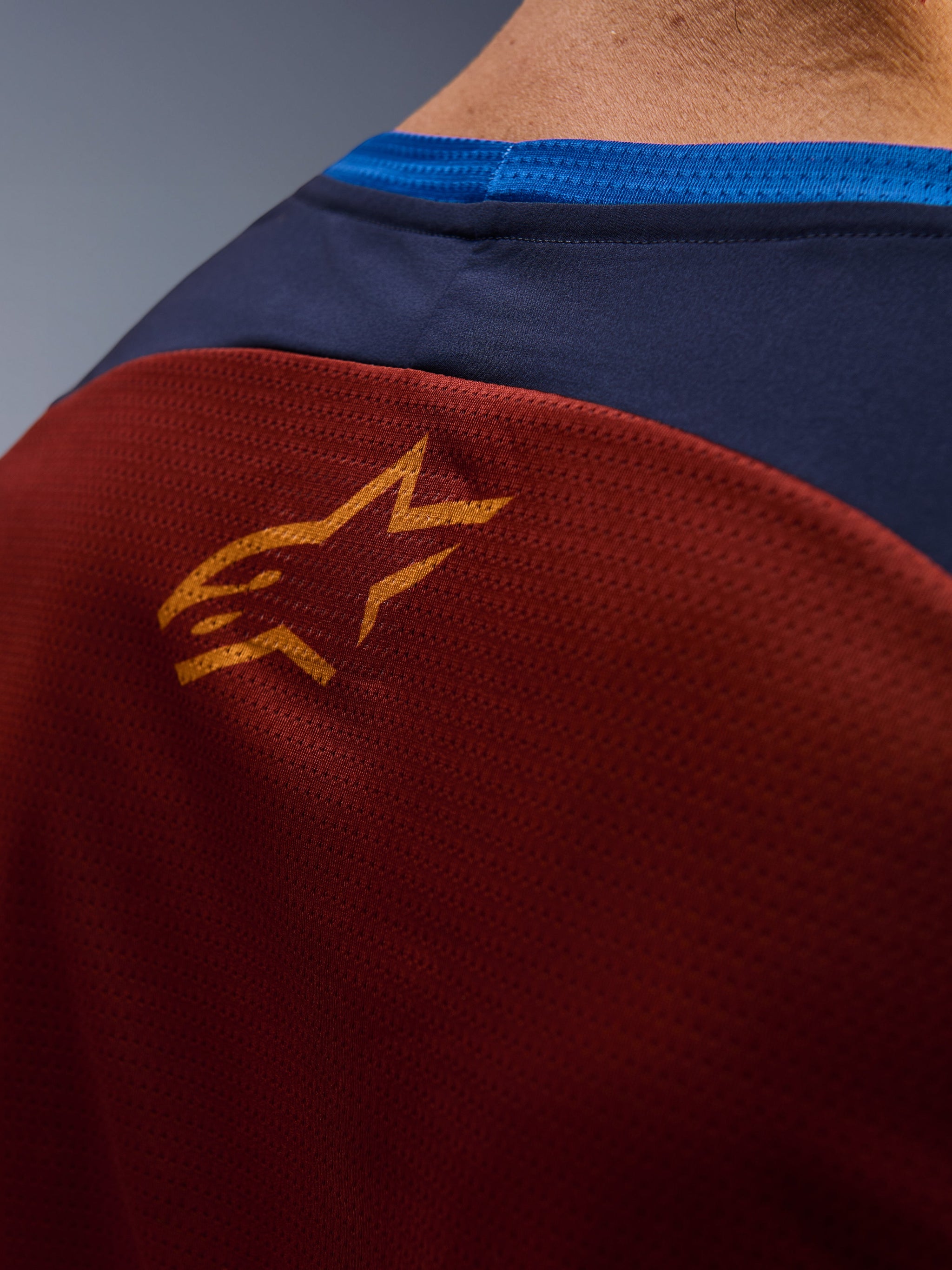 Close-up of Alpinestars Maxdura Dual Jersey, Rust Brown mesh fabric on the back, small orange star logo, dark blue upper panel and bright blue collar trim, detailed view of adventure motorcycle jersey