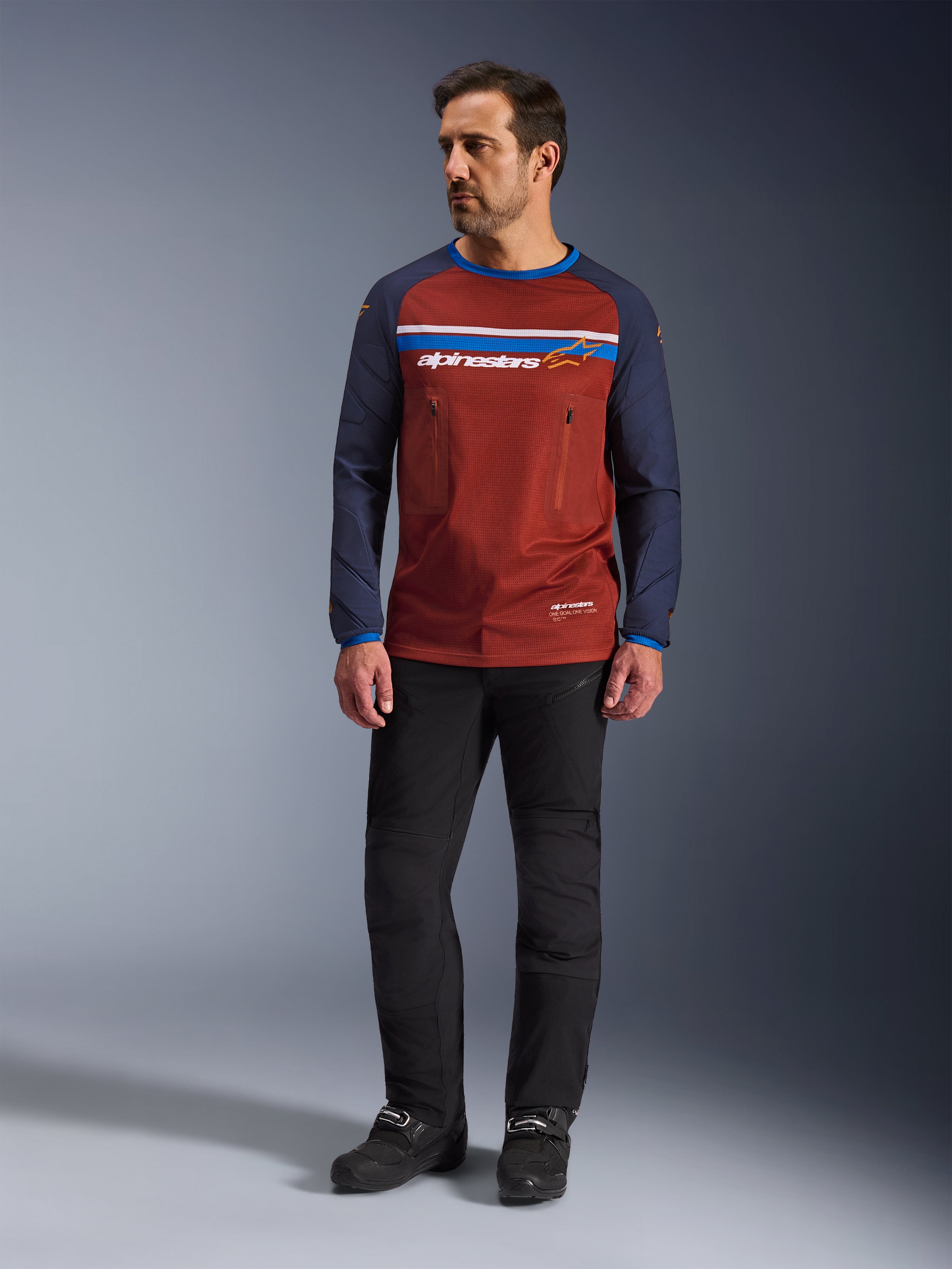 Alpinestars Maxdura Dual Jersey, Rust Brown en Mood Indigo, adventure motorjersey met een mesh-torso en navy mouwen, gedragen door een model met zwarte rijbroek en zwarte laarzen, technische dual-sport kleding voor off-road rijden