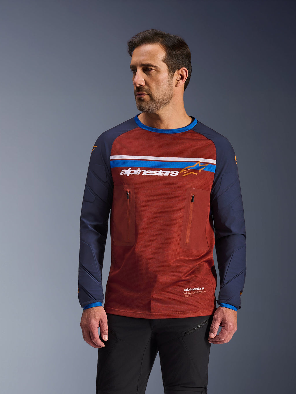 Alpinestars Maxdura Dual Jersey, mannelijk model draagt roestbruine en marineblauwe adventure motorjersey, voorzien van dubbele borstzakken met ritssluiting, horizontale borststreep met logo, technische mesh-torso en duurzame geweven mouwen