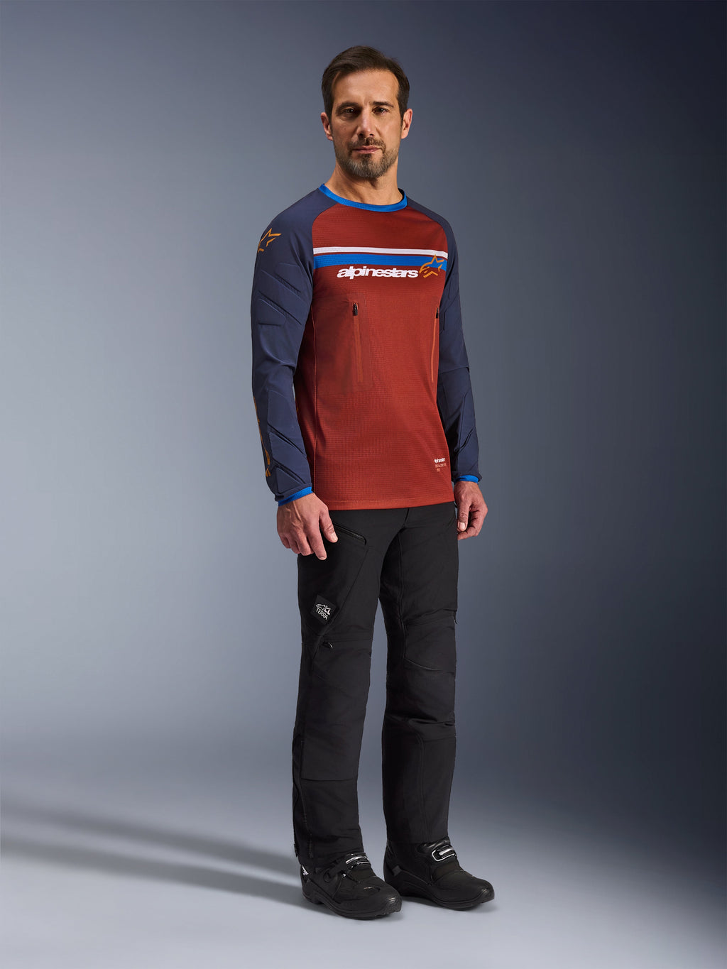 Man draagt de Alpinestars Maxdura Dual Jersey in Rust Brown en Mood Indigo, met een mesh torso en gewatteerde mouwen, gecombineerd met een zwarte motorbroek en laarzen, adventure motorkleding.