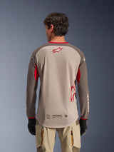 Achteraanzicht van een man die het Alpinestars Maxdura Dual Jersey in Aluminum Walnut draagt, adventure motorjersey met een beige mesh romp en rood sterlogo, donkerbruine mouwen met rode accenten, gecombineerd met een beige motorbroek en zwarte handschoenen, ontworpen voor hoogwaardige off-road ventilatie.