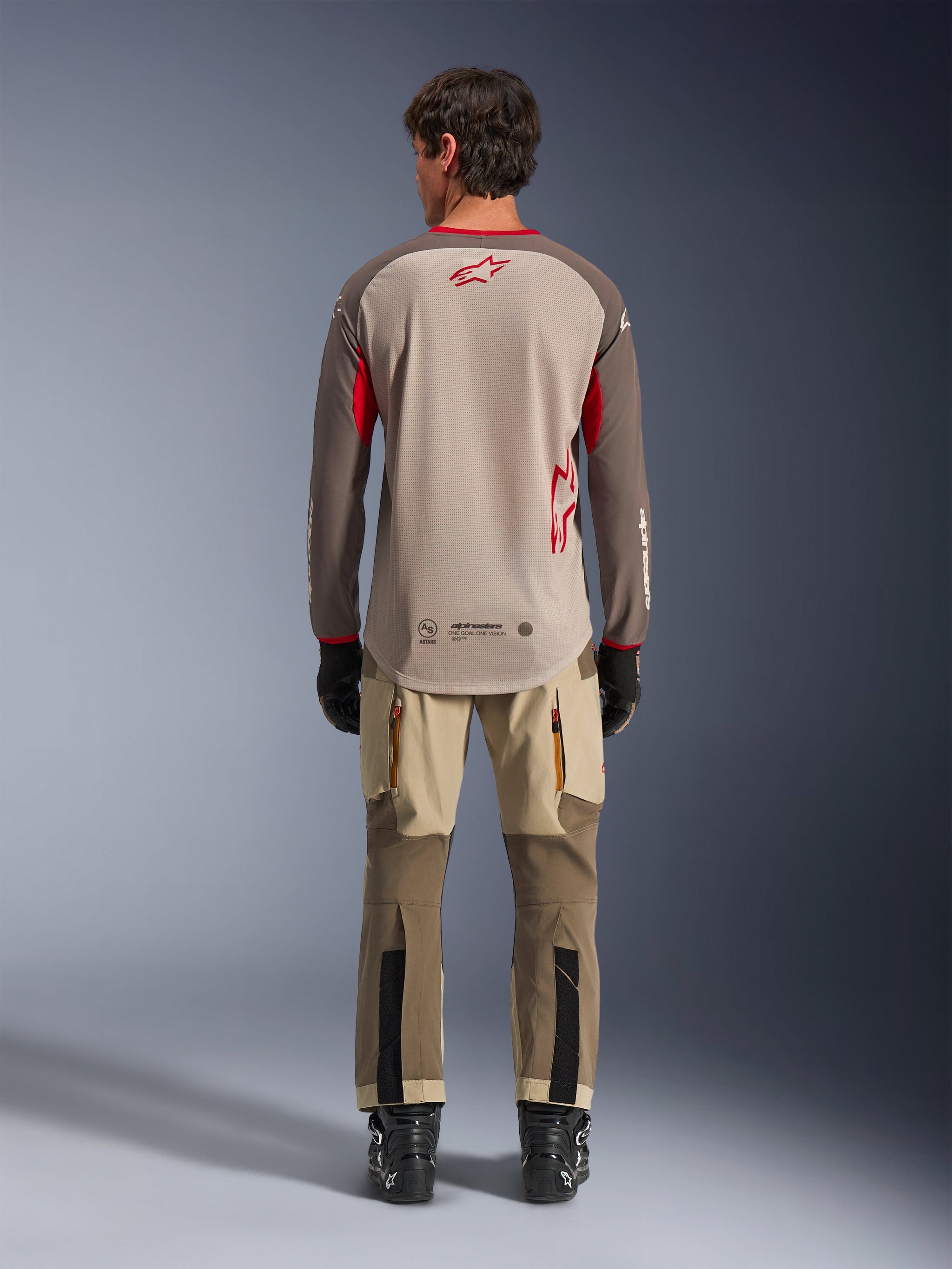 Alpinestars Maxdura Dual Jersey in Aluminum Walnut, achteraanzicht, beige mesh torso met bruine mouwen en rode logo's, verlaagde achterzoom, gecombineerd met adventure motorbroek en zwarte laarzen.