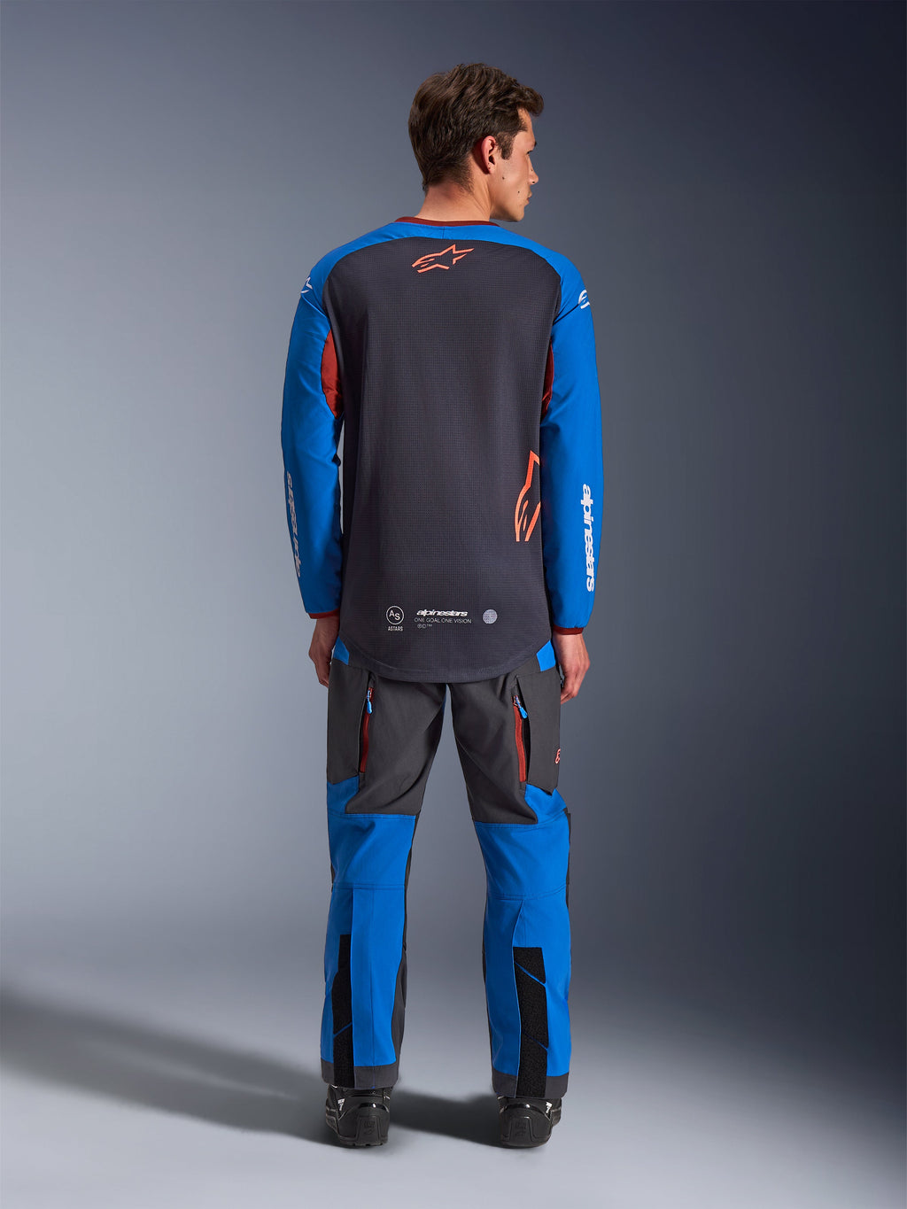 Alpinestars Maxdura Dual Jersey in Deep Gray en Royal Blue, achteraanzicht met volledige mesh torso en oranje sterlogo, koningsblauwe mouwen met witte branding, gedragen met bijpassende grijze en blauwe adventure-broek, hoogwaardige uitrusting voor dual-sport ritten